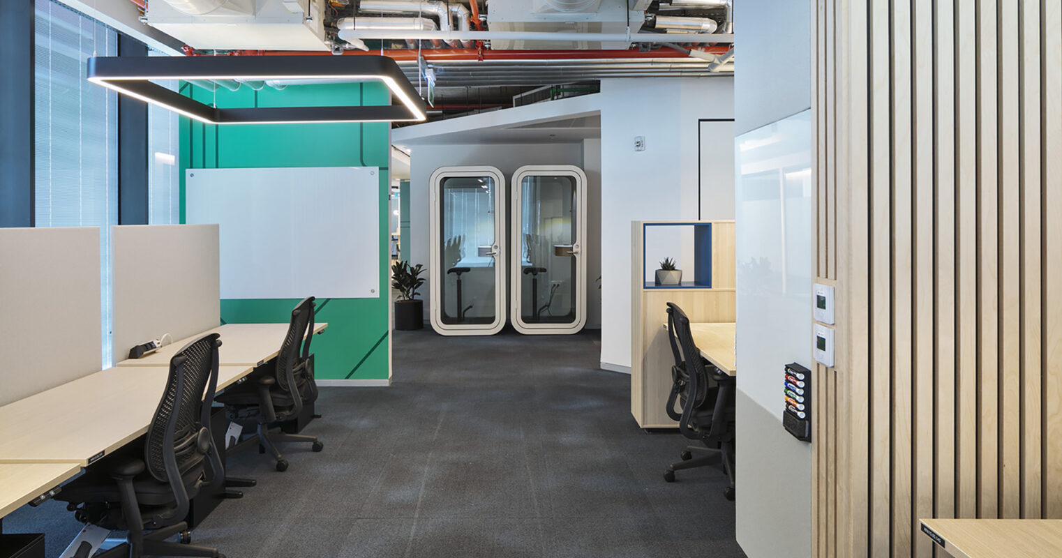 Modern office interior showcasing an open workspace with ergonomic black chairs and light wood desks. A vibrant green accent wall adds a pop of color, complemented by sleek, glass-enclosed meeting rooms. The space features a contemporary design with strategic lighting and sound-absorbing partitions to enhance productivity.