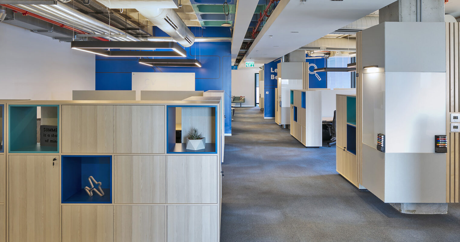 Modern office space featuring modular cubicles with wood paneling and vibrant blue accents. The design prioritizes collaboration and privacy, with strategically placed glass panels offering transparency while maintaining a sense of enclosure. Innovative lighting and an open layout enhance productivity and create a welcoming atmosphere.