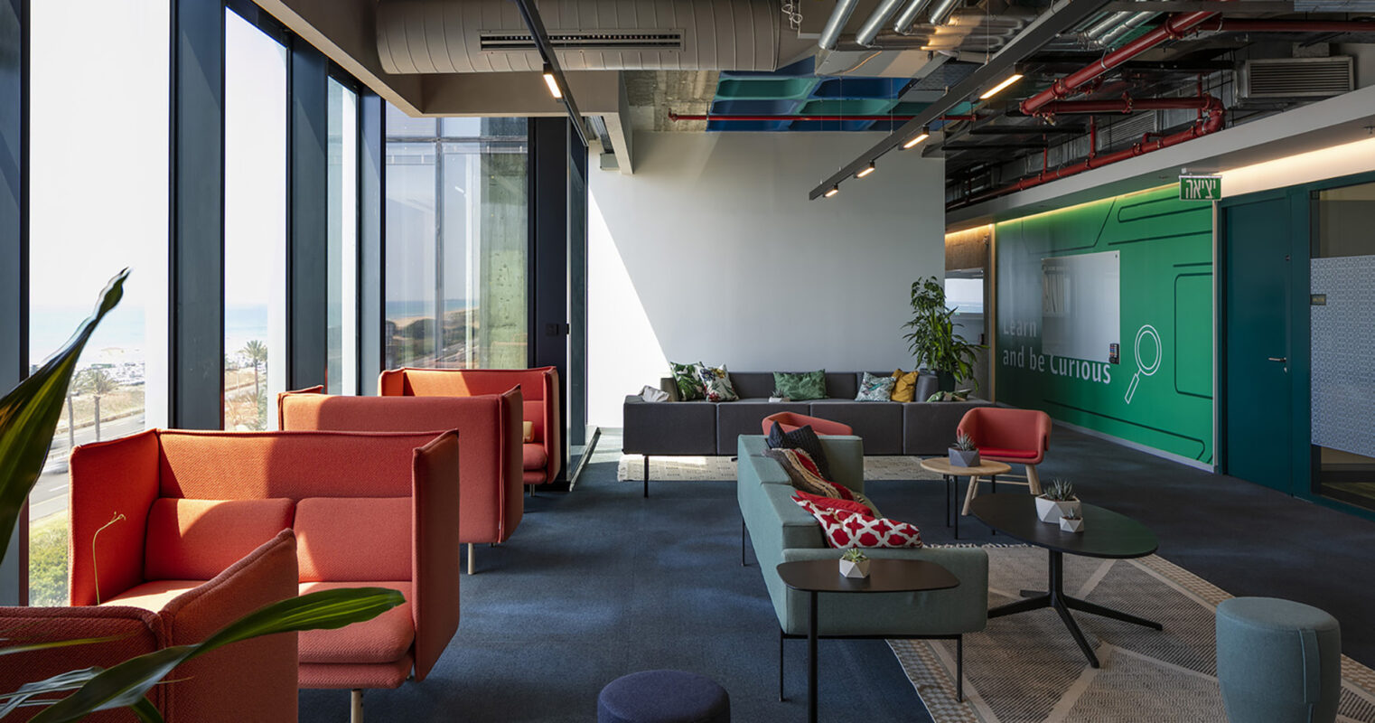 Brightly colored seating arrangements create a vibrant lounge area, featuring modern, angular furniture that encourages collaboration. Expansive windows allow natural light to flood the space, emphasizing the contrast between the rich blue carpet and bold upholstery. Greenery adds an organic touch, enhancing the inviting atmosphere of the contemporary office.