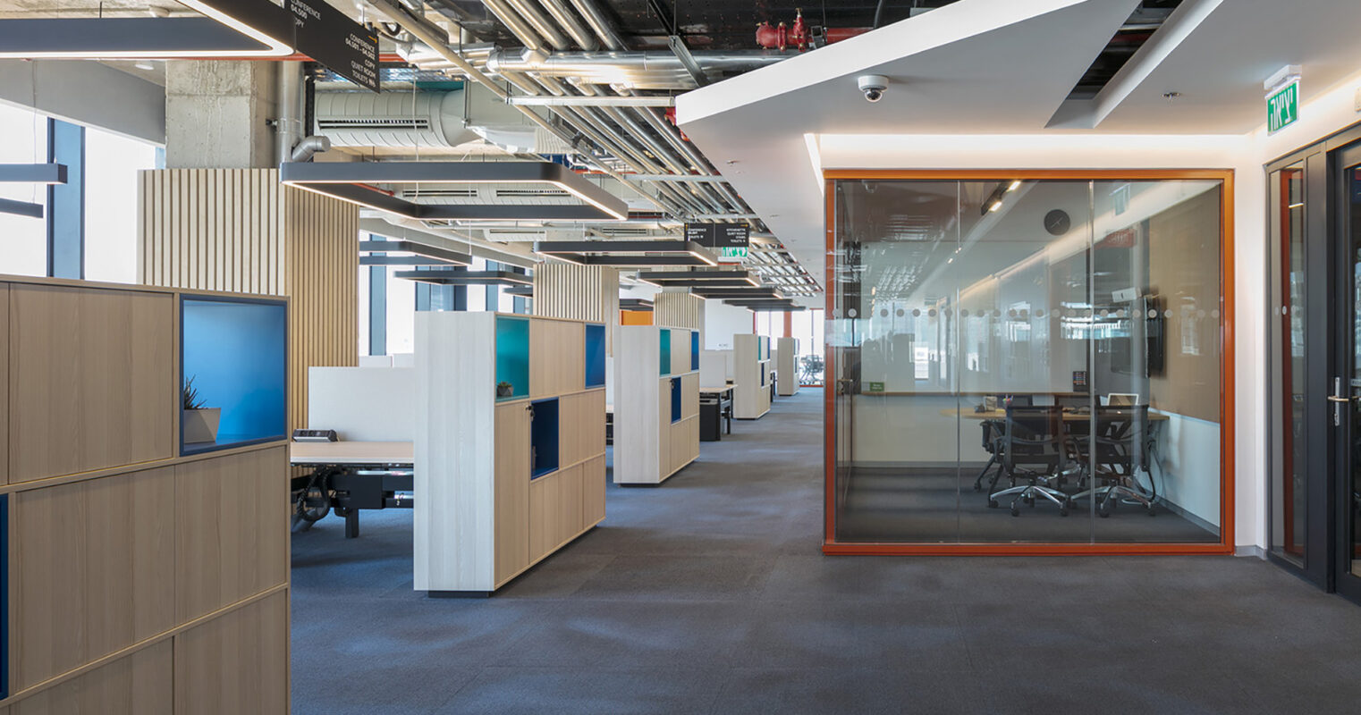 Contemporary office space featuring open-plan workstations divided by modern wooden screens with integrated storage and colorful panels. Natural light floods the area through large windows, while a glass-walled conference room offers a sleek, private space. Exposed ductwork and minimalist design elements enhance the industrial aesthetic.
