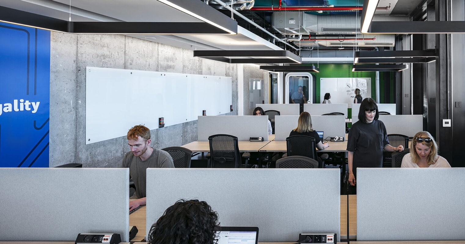Bright, open office space featuring modern workstations with low partitions for privacy. Natural light floods through large windows, enhancing the neutral palette of concrete and wood. Sleek, curved light fixtures provide illumination, while collaboration areas are defined by vibrant wall graphics, promoting a dynamic and innovative work environment.