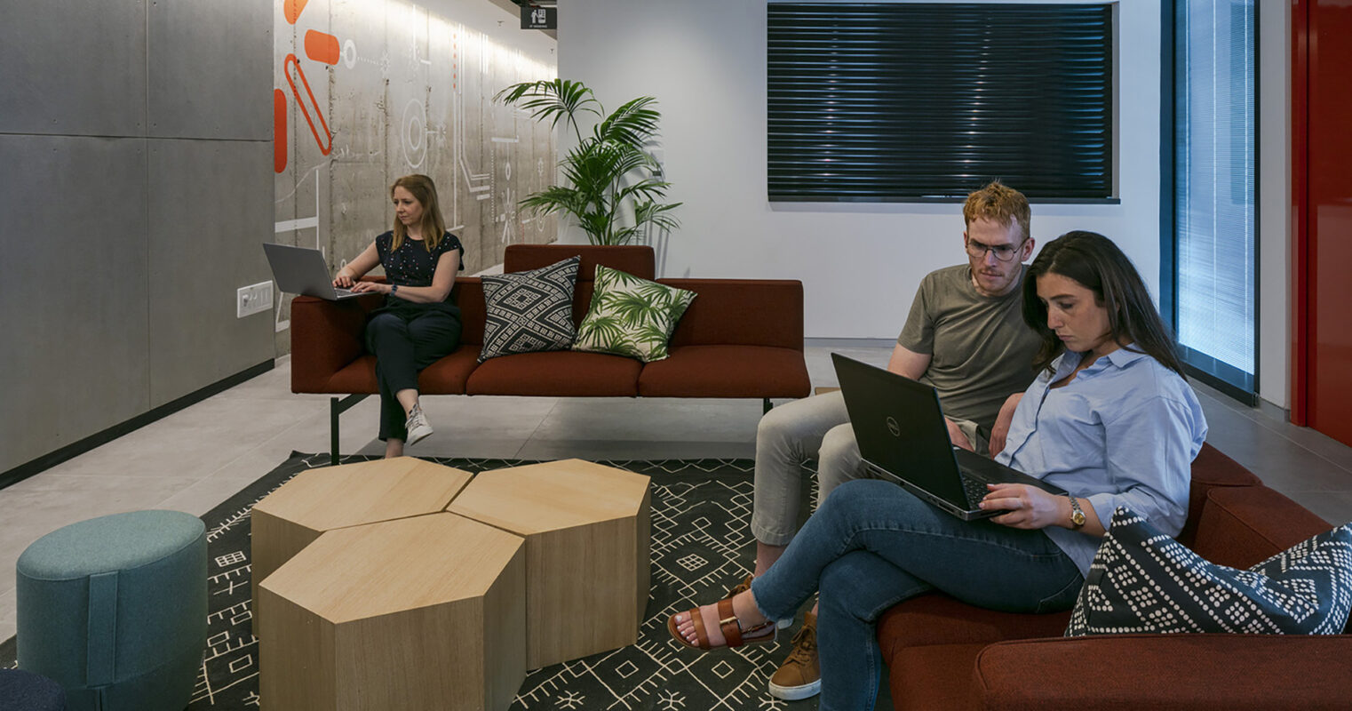 A modern collaborative workspace featuring a contemporary sectional sofa in rich burgundy, complemented by geometric-patterned cushions. A wooden hexagonal coffee table anchors the space, surrounded by stylish poufs in varying shades. Natural light streams in through large windows, enhancing the inviting ambiance for productive engagement.