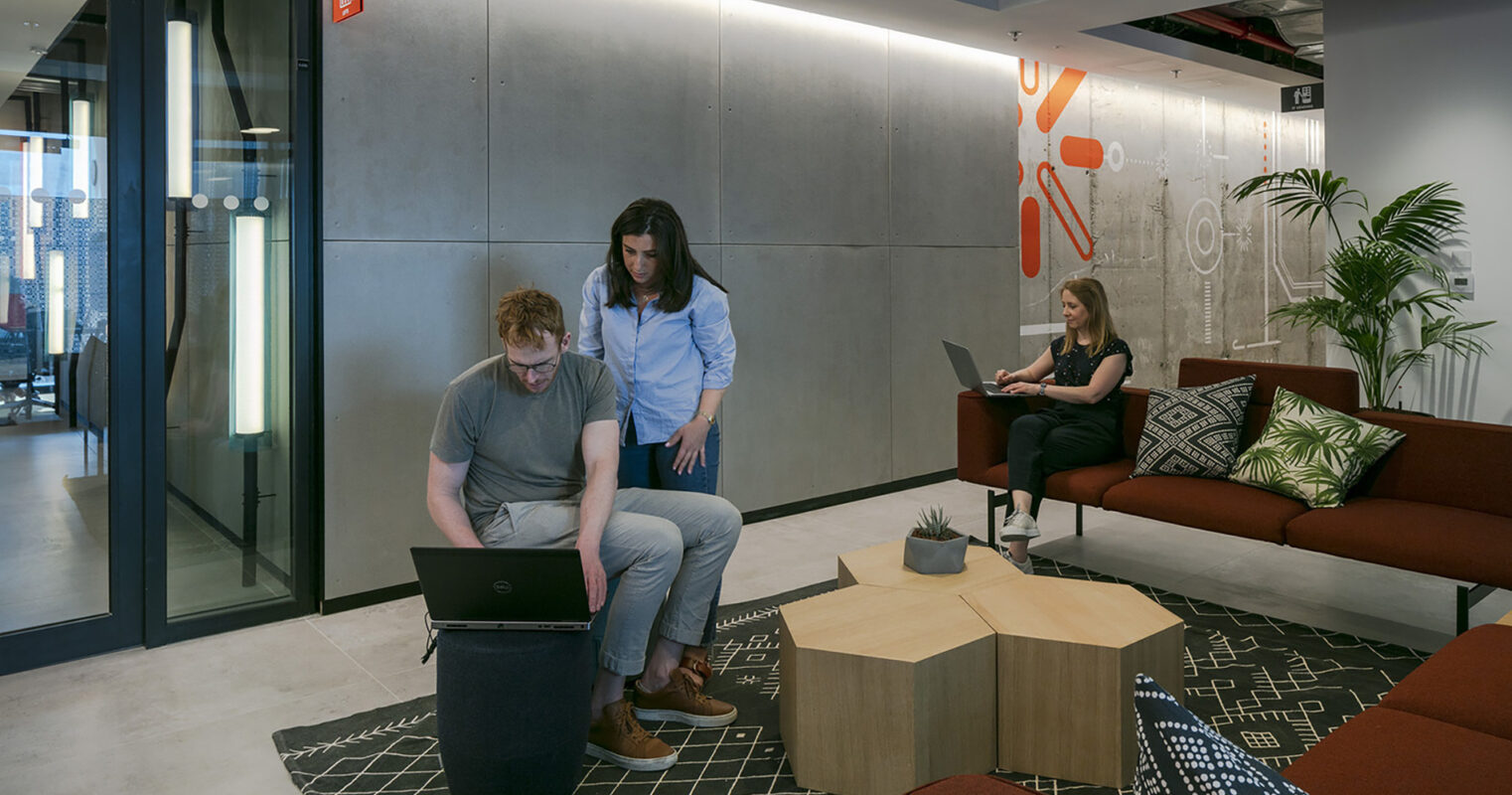 Collaborative workspace featuring a modern design aesthetic. A man sits on a cylindrical seat, reviewing a laptop with a woman standing beside him. In the background, a woman works on her laptop, surrounded by contemporary furnishings, including a geometric coffee table and a patterned rug, fostering a productive environment.