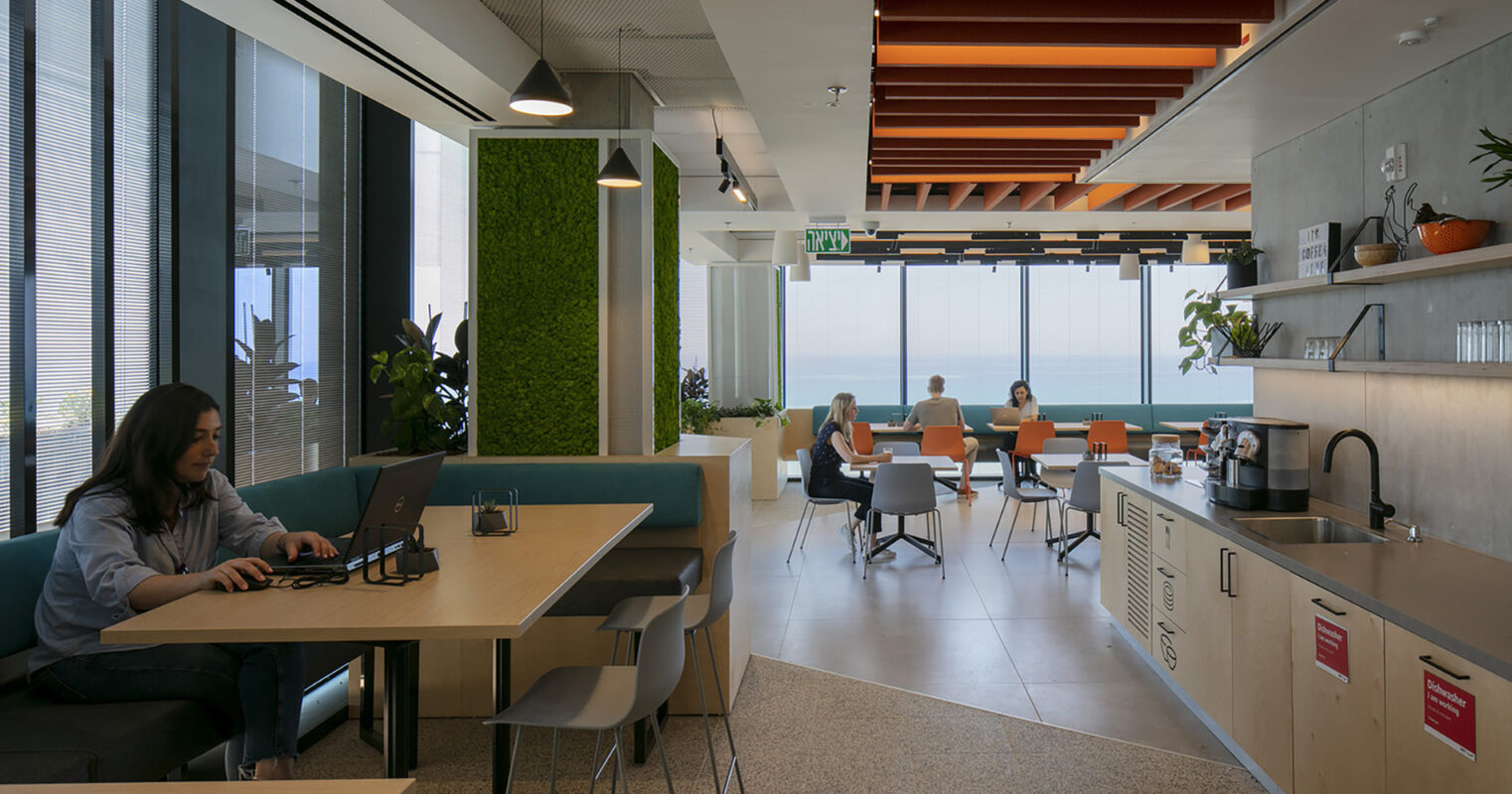 A modern workspace showcases an open layout with large windows flooding the area with natural light. Stylish seating arrangements include a cozy nook with a laptop user and a communal table where colleagues engage. Biophilic design elements like green wall accents enhance the ambiance, promoting creativity and collaboration.