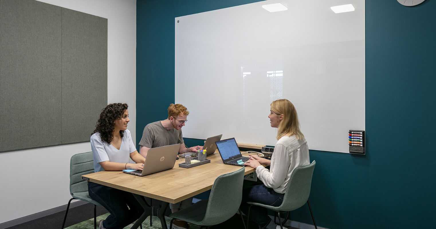 A collaborative workspace features three individuals engaged in a meeting around a minimalist wooden table. The walls are painted a rich teal, complemented by a large whiteboard for brainstorming. Soft acoustic panels enhance sound absorption, while a textured green rug adds warmth and comfort to the modern aesthetic.