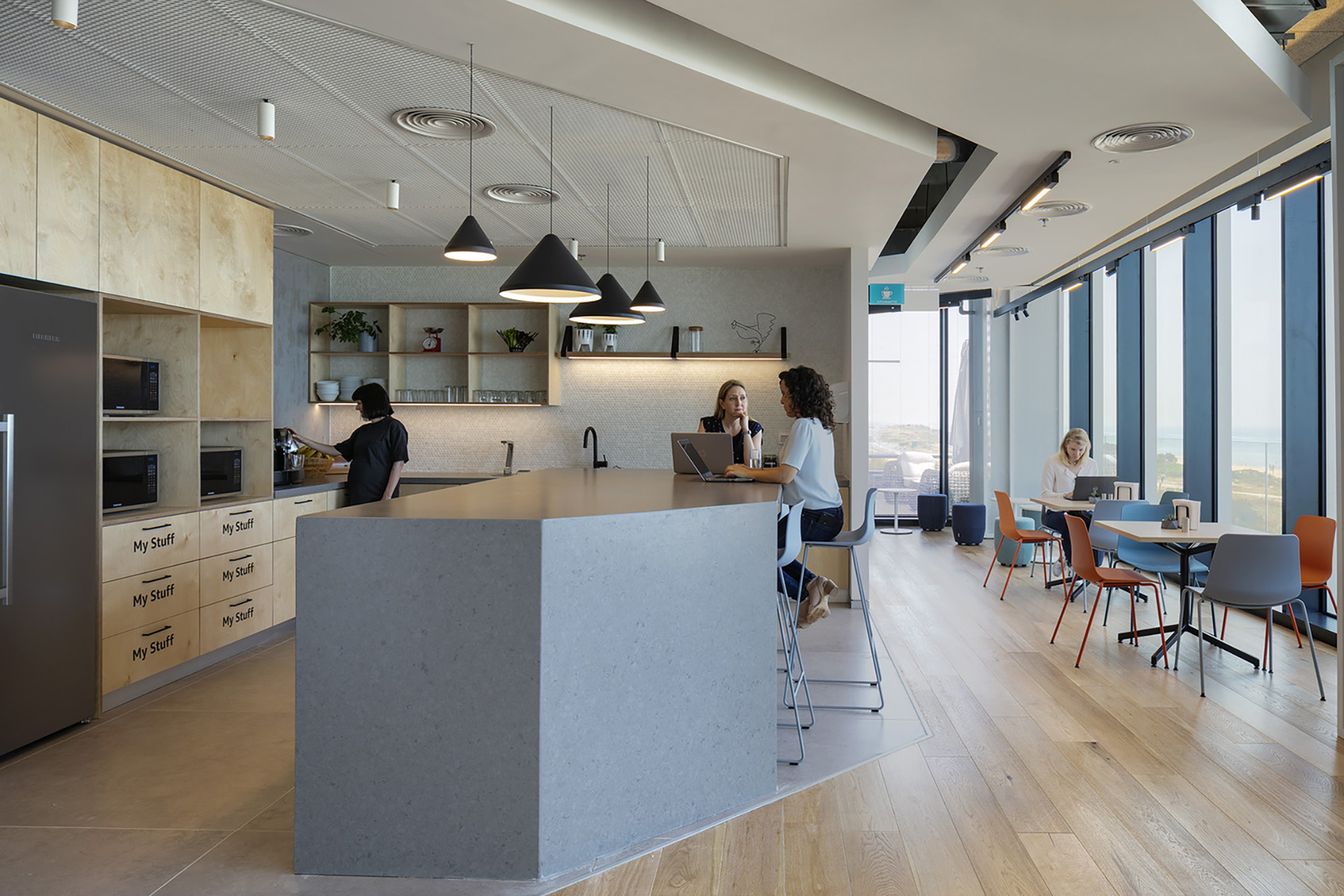 Modern open-plan kitchen featuring a large central island in grey stone, complemented by wooden cabinetry. Pendant lights create a warm ambiance. Three individuals engage in conversation, while a fourth person works on a laptop at a nearby table. Floor-to-ceiling windows showcase scenic views, enhancing the space's natural light.