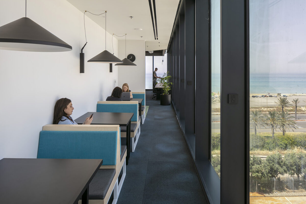 Modern workspaces feature sleek, minimalist booths with turquoise upholstery, designed for comfort and collaboration. Large windows offer panoramic views of the ocean, enhancing the serene atmosphere. Pendant lighting complements the design, while individuals engaged in work create a vibrant, productive environment, merging functionality with aesthetic appeal.