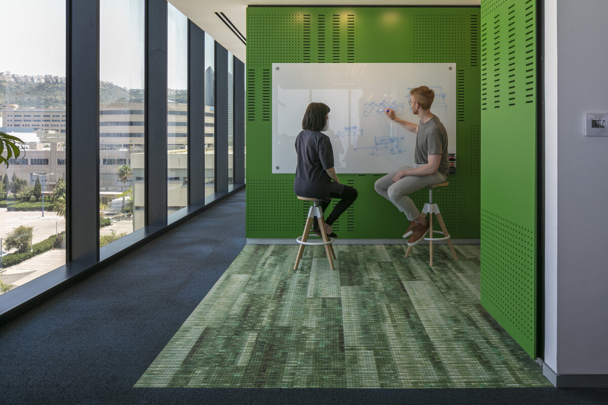 Modern office interior featuring a vibrant green accent wall with perforated panels, promoting collaboration. Two professionals engage in discussion while seated on bar stools, with a whiteboard displaying sketches to their left. The floor combines textured carpet and patterned tiles, enhancing the aesthetic appeal and functional design.