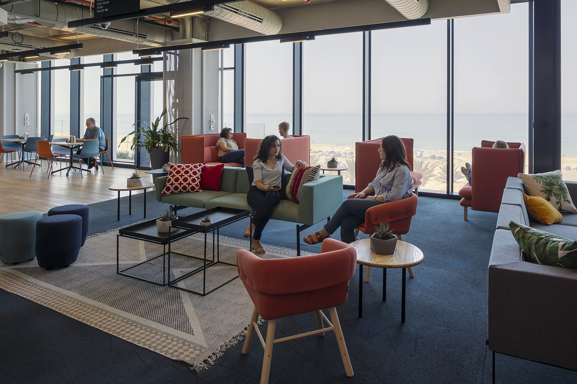 Contemporary lounge area featuring a mix of vibrant textile patterns and comfortable seating options, including modular sofas and armchairs. Large windows frame a panoramic view of the ocean, enhancing natural light. Indoor plants add a touch of greenery, creating an inviting atmosphere for collaboration and relaxation.