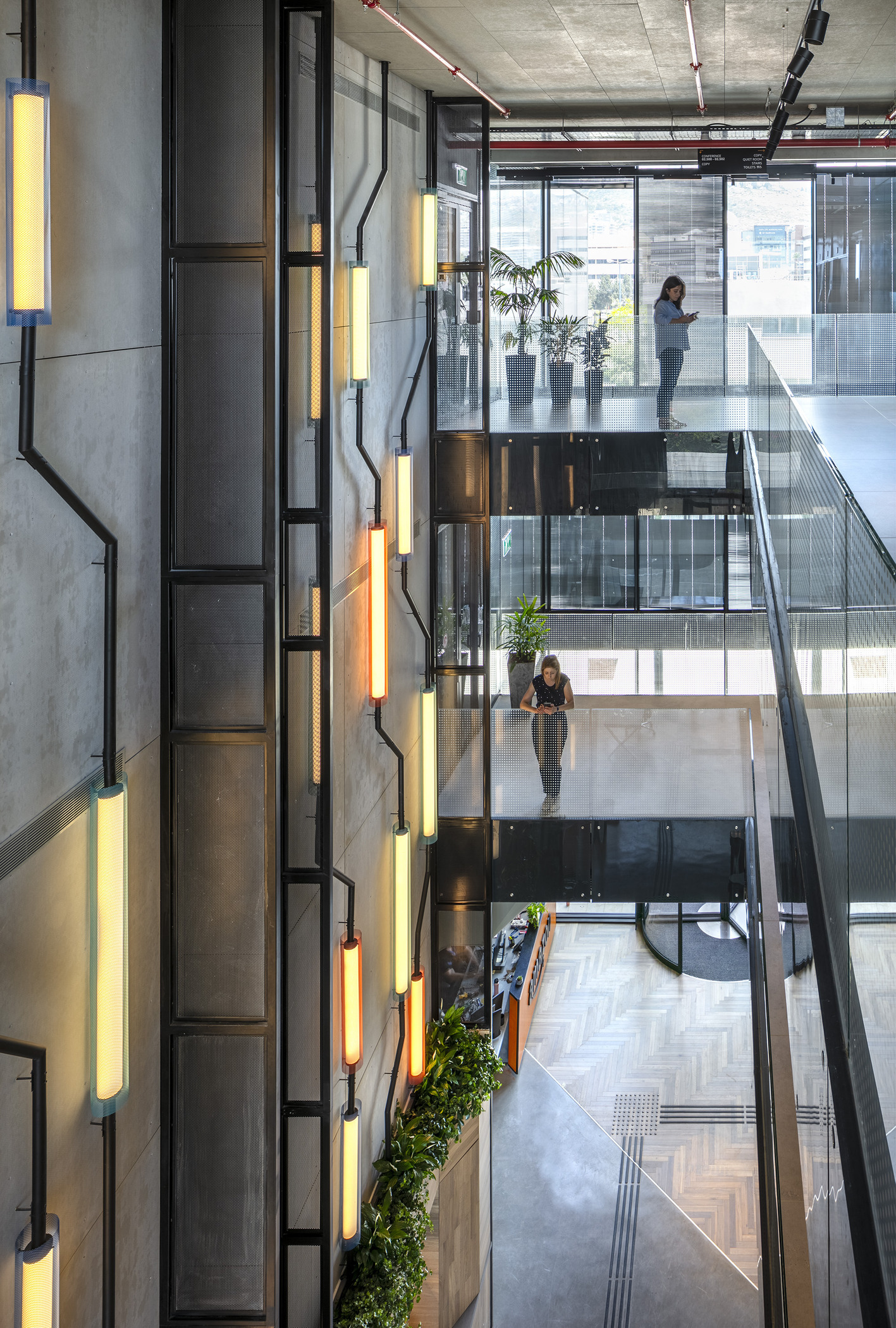 A modern multi-story atrium featuring sleek concrete walls and industrial design elements. Colorful wall-mounted light fixtures illuminate the space, while glass railings provide open sightlines. Potted plants add a touch of greenery, contrasting with the otherwise minimalist aesthetic. The layout promotes a sense of openness and collaboration.