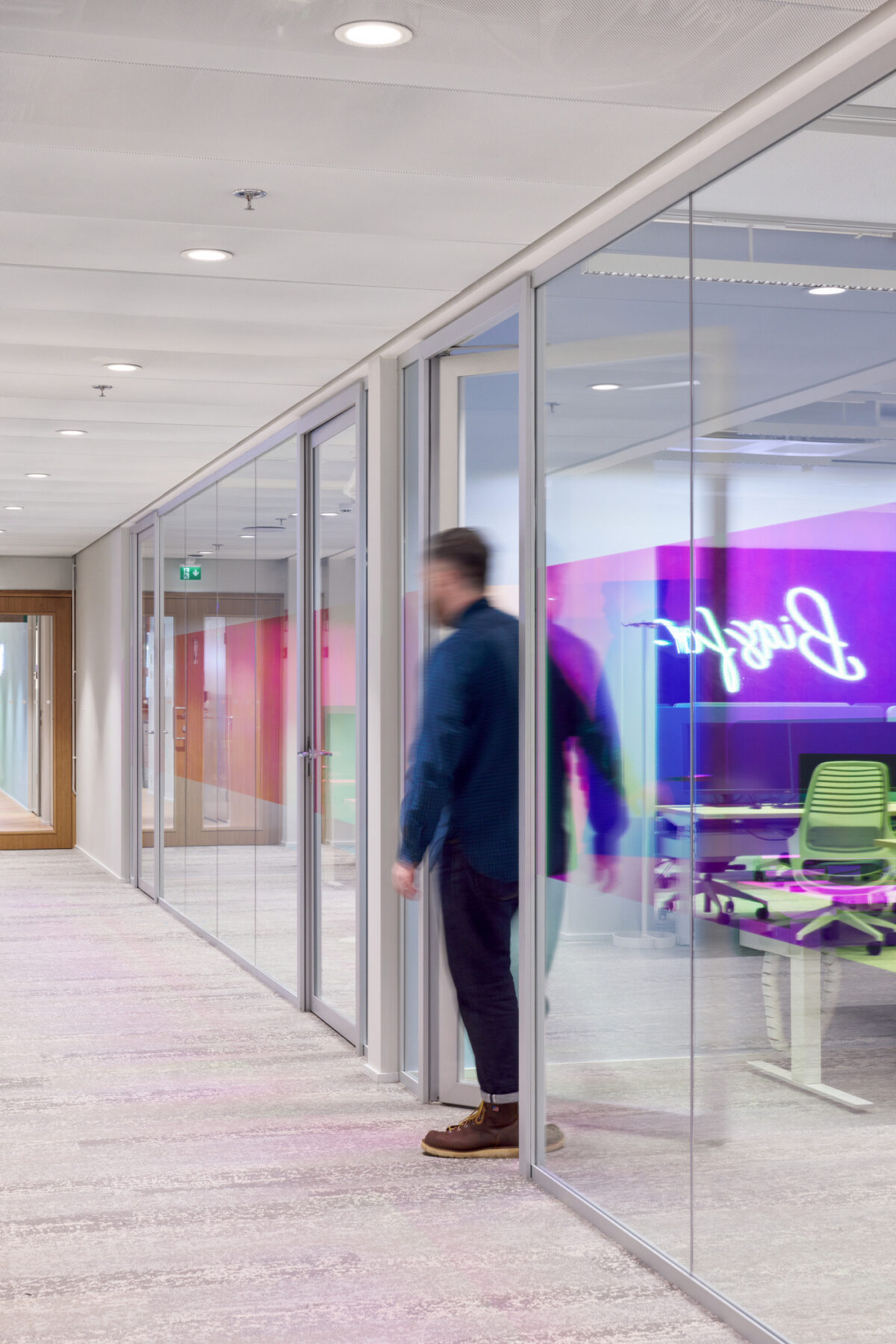 Brightly lit corridor featuring sleek, transparent glass walls that connect modern office spaces. A blurred figure walking past reflects the open layout and collaborative atmosphere. Colorful abstract wall art adds vibrancy, complementing ergonomic furniture in the workspace, promoting creativity and dynamic interaction among employees.