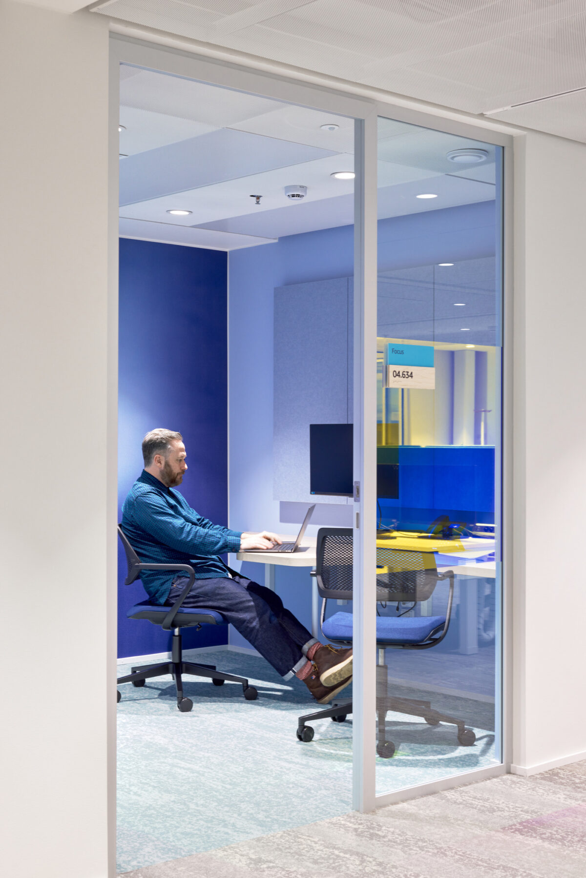 A modern workspace features a man seated at a desk, engrossed in his laptop. The glass walls enhance visibility while offering acoustic privacy. Vibrant blue accents and soft furnishings create a calming environment, showcasing a blend of functionality and contemporary design within an open office concept.
