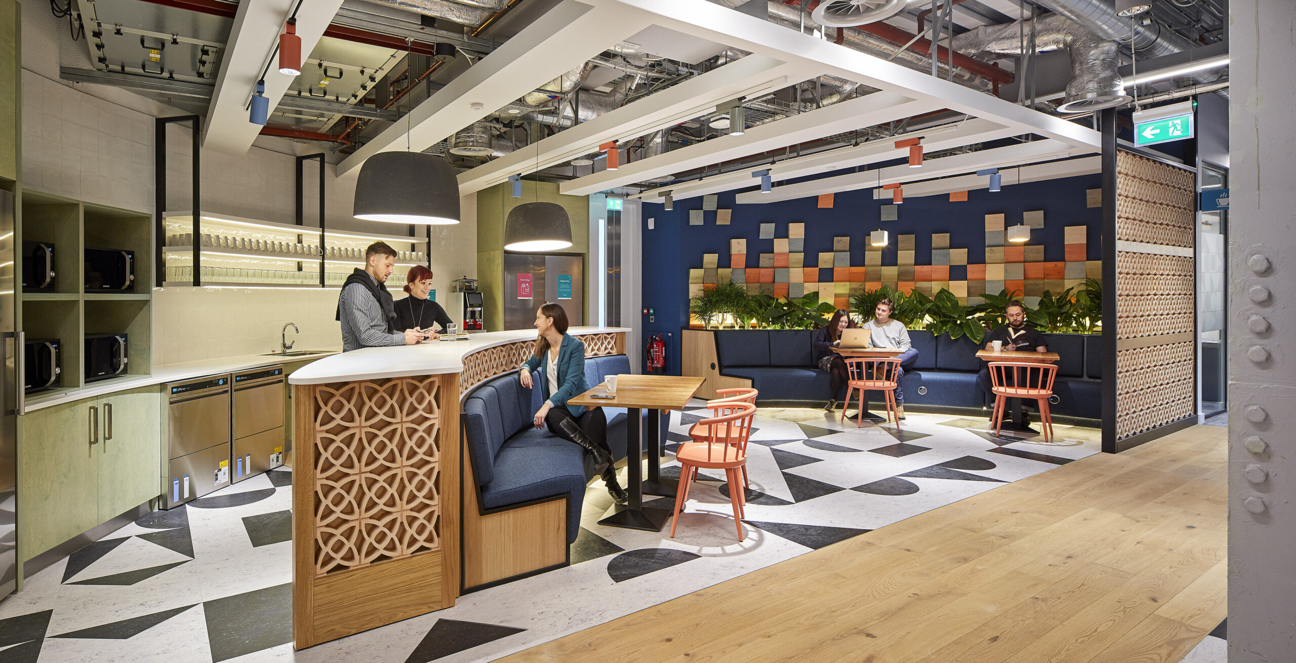 A contemporary coworking space featuring a mix of vibrant colors and textures. The communal area includes geometric black-and-white flooring, wooden accents, and patterned walls. Comfortable seating areas and a modern kitchenette enhance the space's functionality, inviting collaboration and relaxation among professionals. Lush greenery adds a refreshing touch.