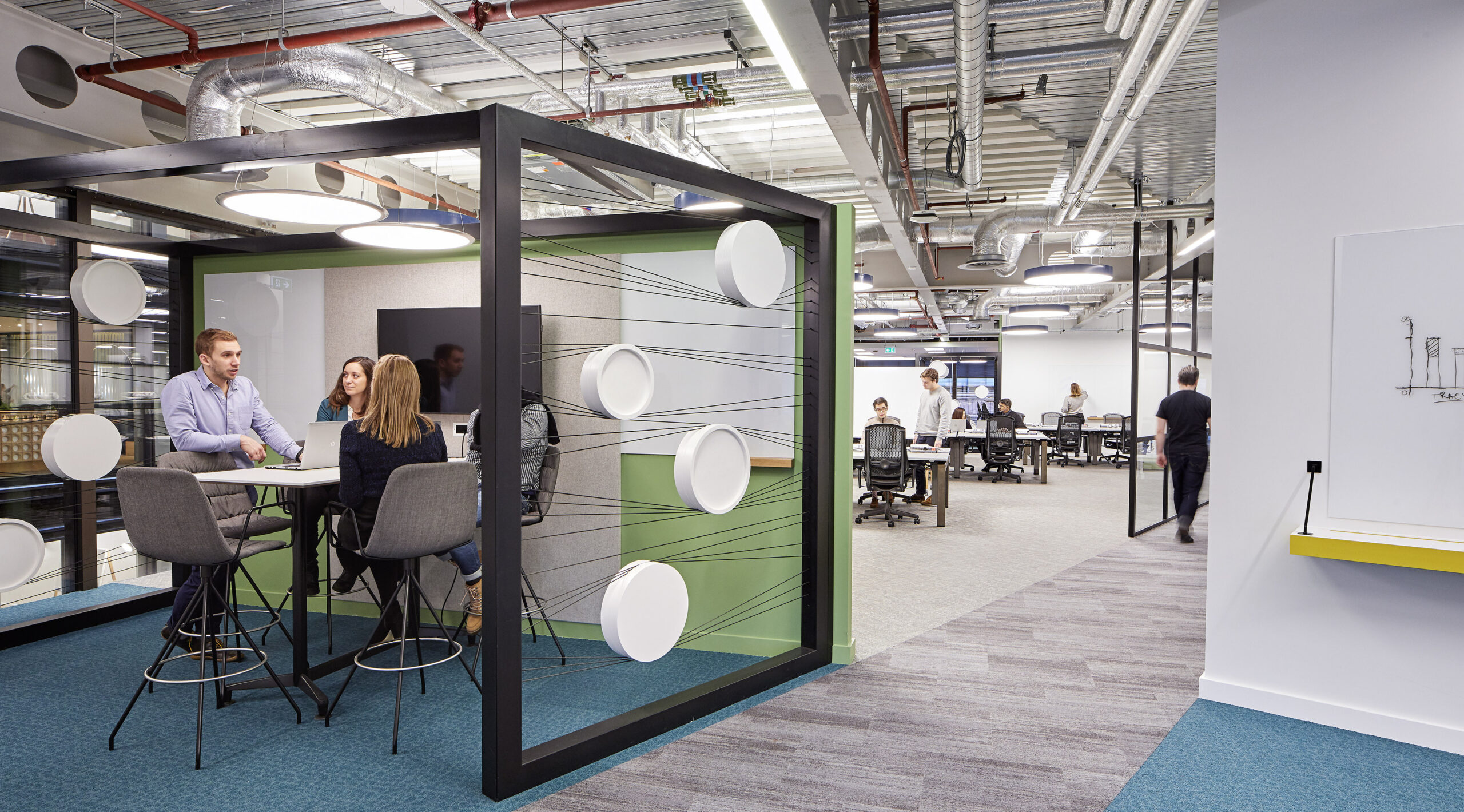A modern office space featuring a collaborative meeting area with a sleek black frame and thin, graphic lines connecting white circular panels. Professionals engage in discussion around a high table, while flexible workstations are visible in the background, emphasizing an adaptable and open design approach.