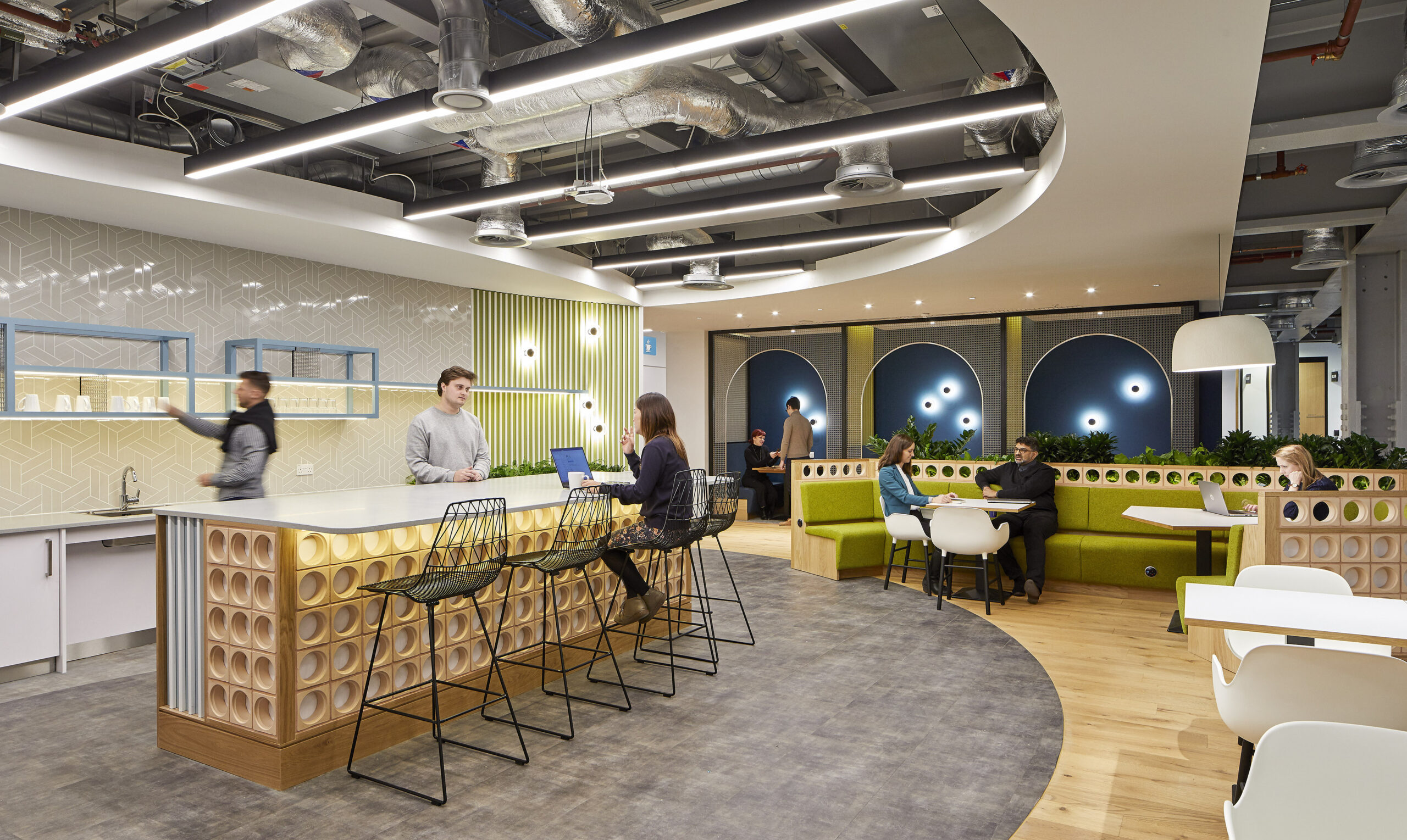 Contemporary office break area featuring a sleek bar with patterned cabinetry and bar stools. The space blends comfortable seating with green accents, ambient circular lighting, and unique wall designs. Engaged individuals collaborate, highlighting the design’s focus on functionality, comfort, and modern aesthetics in a professional environment.