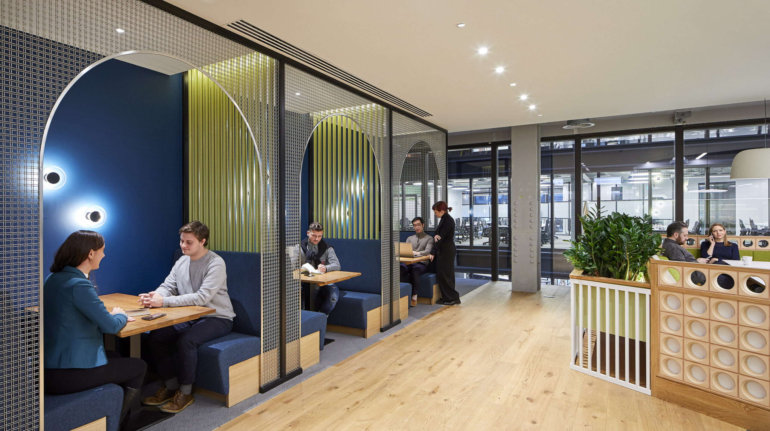 Stylish collaborative workspace featuring cozy booths with blue and green accents. Arched dividers create semi-private areas, fostering conversation and productivity. Warm wood flooring complements contemporary furnishings, while large windows provide ample natural light, enhancing the inviting atmosphere of the modern office environment.