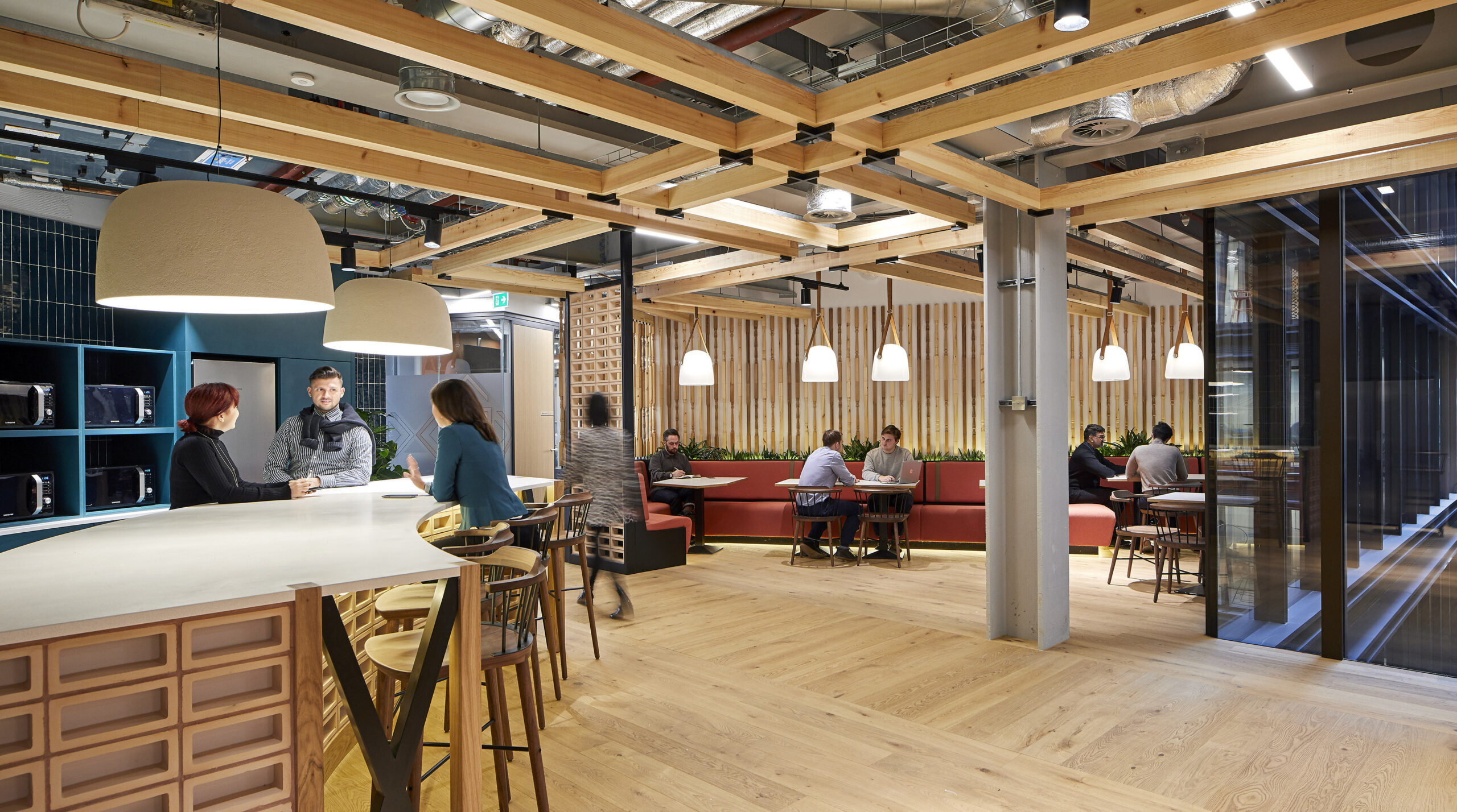 Modern office interior featuring a combination of natural wood and industrial elements. The space includes a large bar-style counter with high stools, pendant lighting, and various seating arrangements. A warm color palette and green plants create a welcoming atmosphere, facilitating collaboration and creativity among employees.
