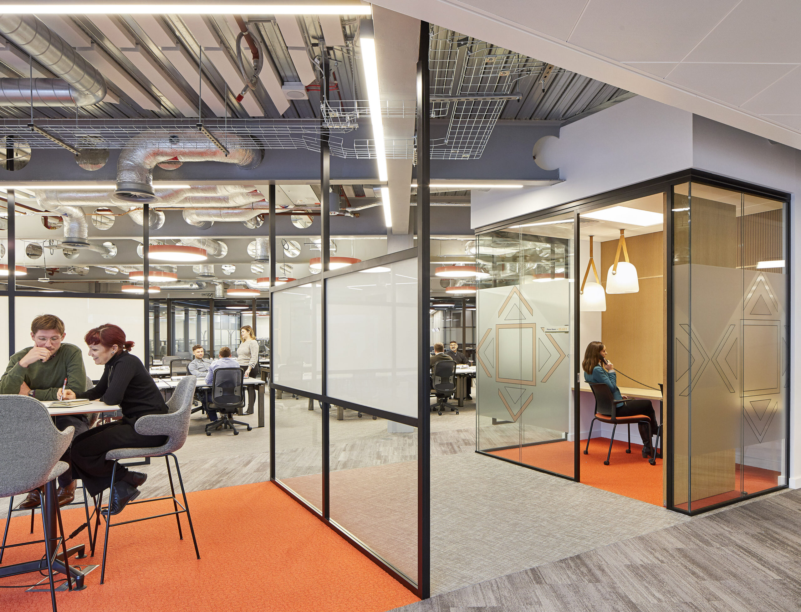Collaborative workspaces feature modern glass partitions, promoting transparency and interaction. The interior combines functional areas with vibrant accents, such as orange carpeting, while overhead lighting enhances the contemporary atmosphere. Private meeting rooms are designed with acoustic glass to ensure privacy without sacrificing light and openness.