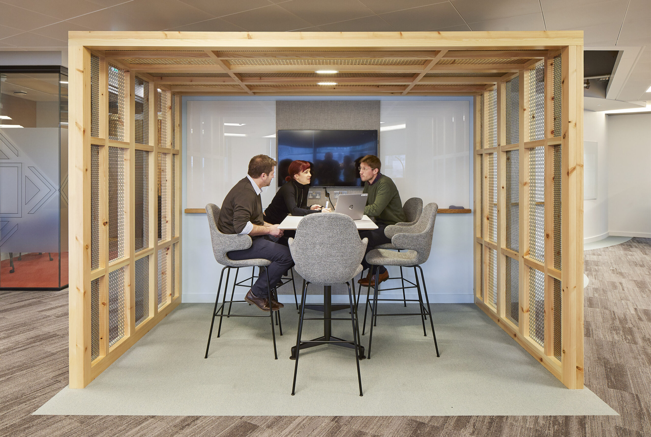 A contemporary collaboration space features a wooden structure with mesh paneling, designed for openness and interaction. Four professionals engage at a circular table, equipped with laptops in a cozy yet modern environment enhanced by soft gray carpet and muted wall colors. The setting promotes teamwork and creativity.
