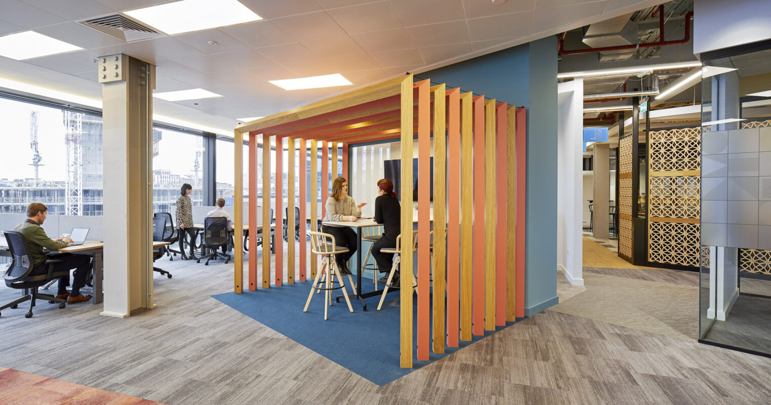 Colorful wooden slats form an open meeting pod within a modern office space, promoting collaboration. Natural light floods the area through large windows, while various workstations and stylish acoustic panels enhance functionality and aesthetics. The design balances privacy and openness, fostering creativity in a professional environment.