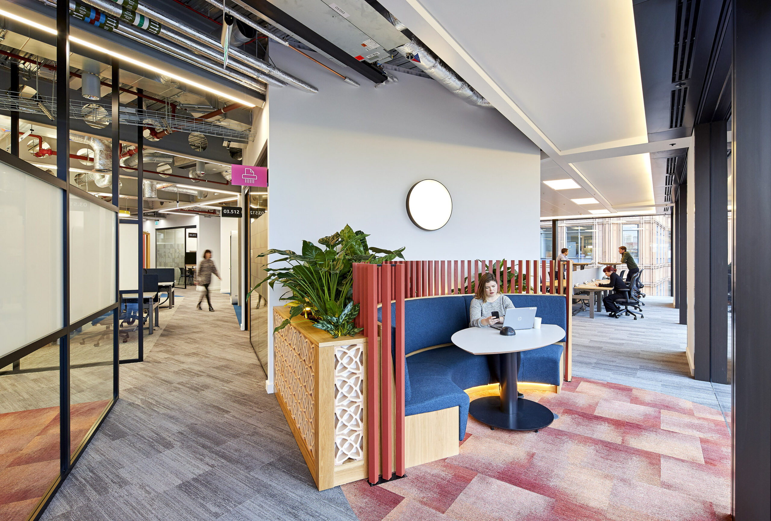 A modern office interior features a cozy collaboration nook, designed with vibrant blue seating and natural wood elements. Lush greenery enhances the space, complemented by an understated circular light fixture. Open workstations are visible in the background, emphasizing a blend of comfort and productivity.