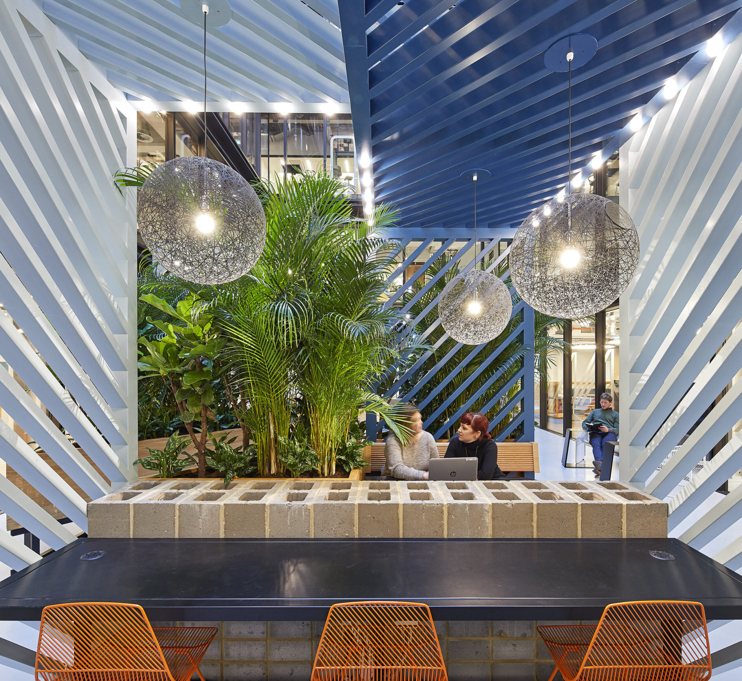 A modern workspace featuring a striking blue and white striped ceiling, complemented by vibrant hanging pendant lights. Lush greenery creates a biophilic atmosphere, while a sleek black table contrasts with textured concrete blocks. Two individuals engage in conversation, enhancing the collaborative and inviting environment.