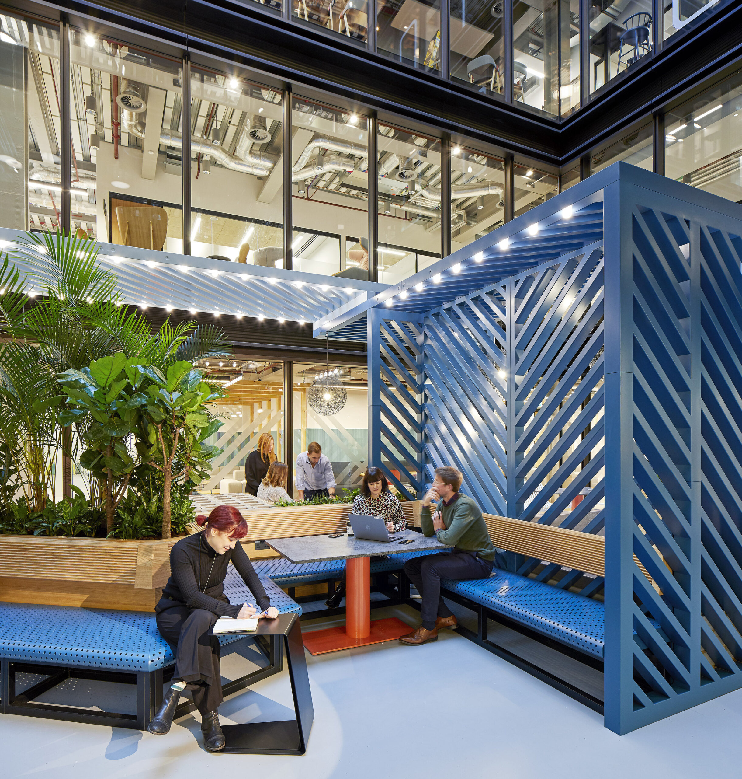 A modern, open communal workspace featuring geometric blue seating and wooden benches, surrounded by lush greenery. The area is illuminated with soft lighting, creating an inviting atmosphere. Employees collaborate and work on laptops, showcasing a blend of comfort and contemporary design in a collaborative environment.