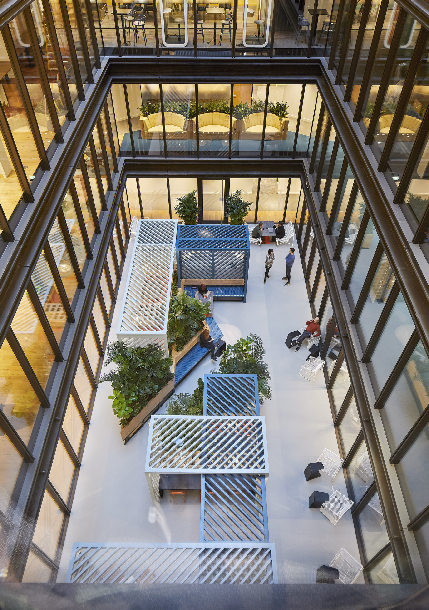 Aerial view of a modern atrium featuring a unique, multi-level layout with slatted wooden elements and integrated greenery. Crisp glass walls enhance natural light, while seating areas are surrounded by vibrant plants, creating a harmonious blend of indoor and outdoor spaces. The design promotes collaboration and tranquility.
