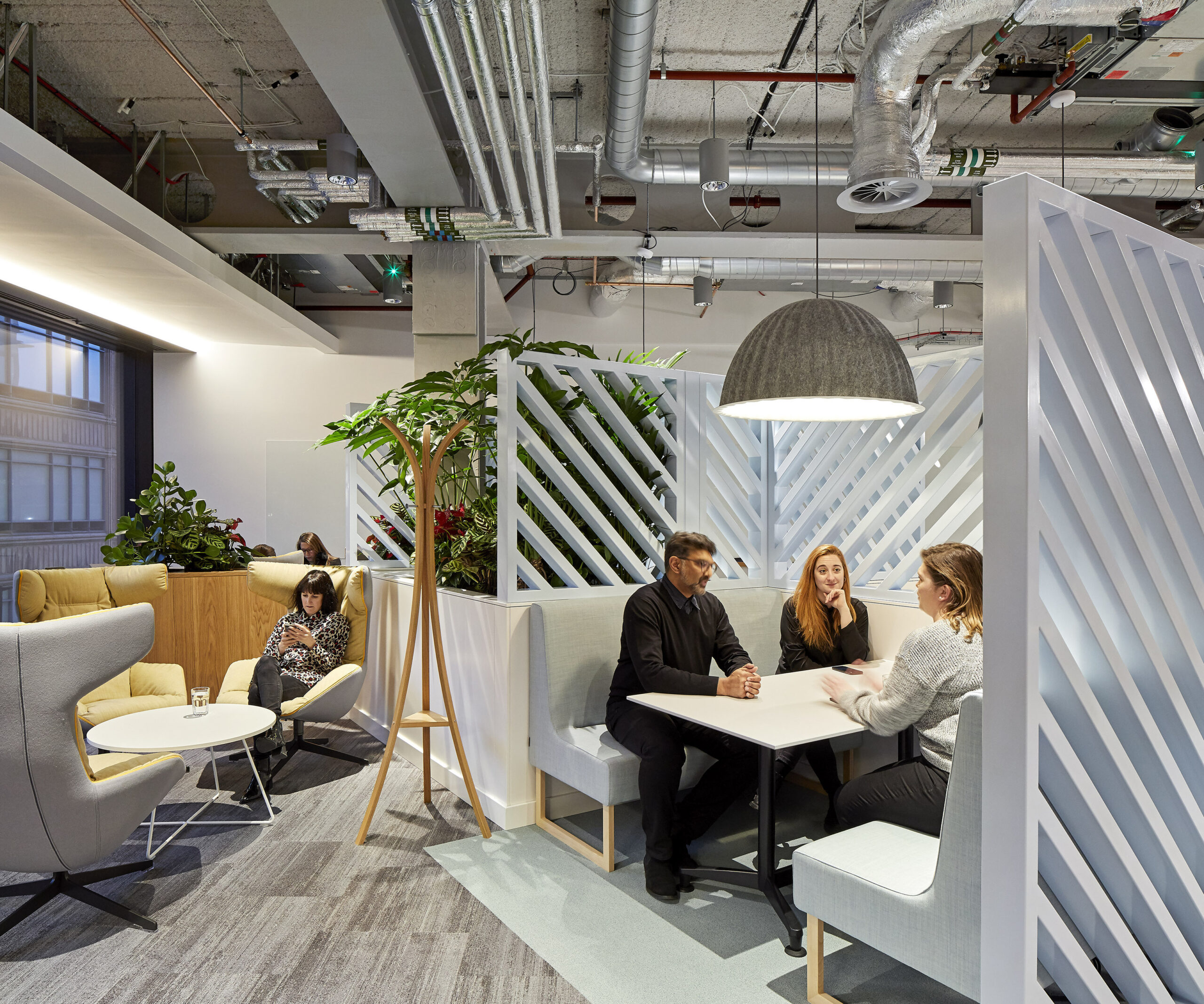 A modern office lounge features stylish booth seating arranged around a light wood table, encouraging collaboration. Lush greenery enhances the space, while a large pendant light adds an industrial touch. Slim, diagonal partitions provide privacy, balancing openness with a contemporary aesthetic that promotes productivity and comfort.