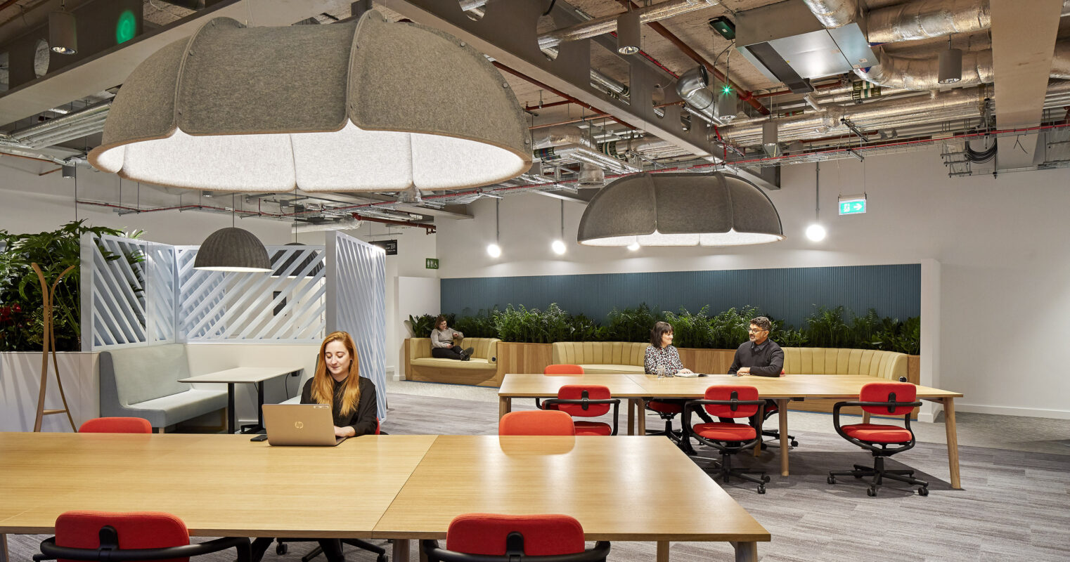 Modern office interior featuring flexible workspaces. A central wooden table with ergonomic red chairs accommodates collaboration. Soft, oversized pendant lights create a warm atmosphere, complemented by greenery and textured partitions. The open layout encourages interaction while maintaining a balance of privacy, demonstrating effective spatial design in a professional environment.