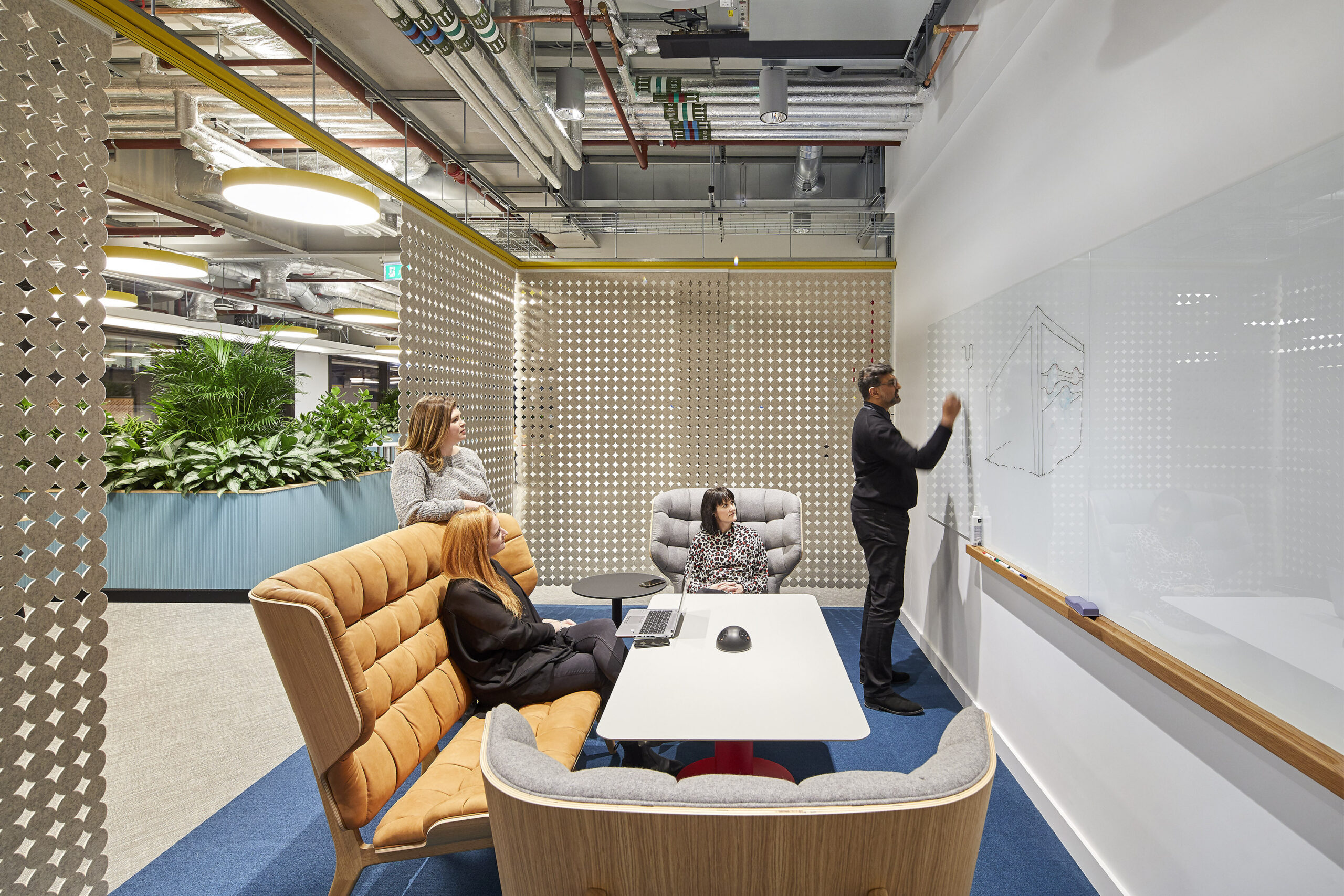 A modern collaborative workspace featuring a cozy seating area with plush armchairs and a central table. A person is drawing on a whiteboard while two individuals observe. The space is accented with greenery in the background and a textured, perforated screen, enhancing both aesthetics and acoustics.