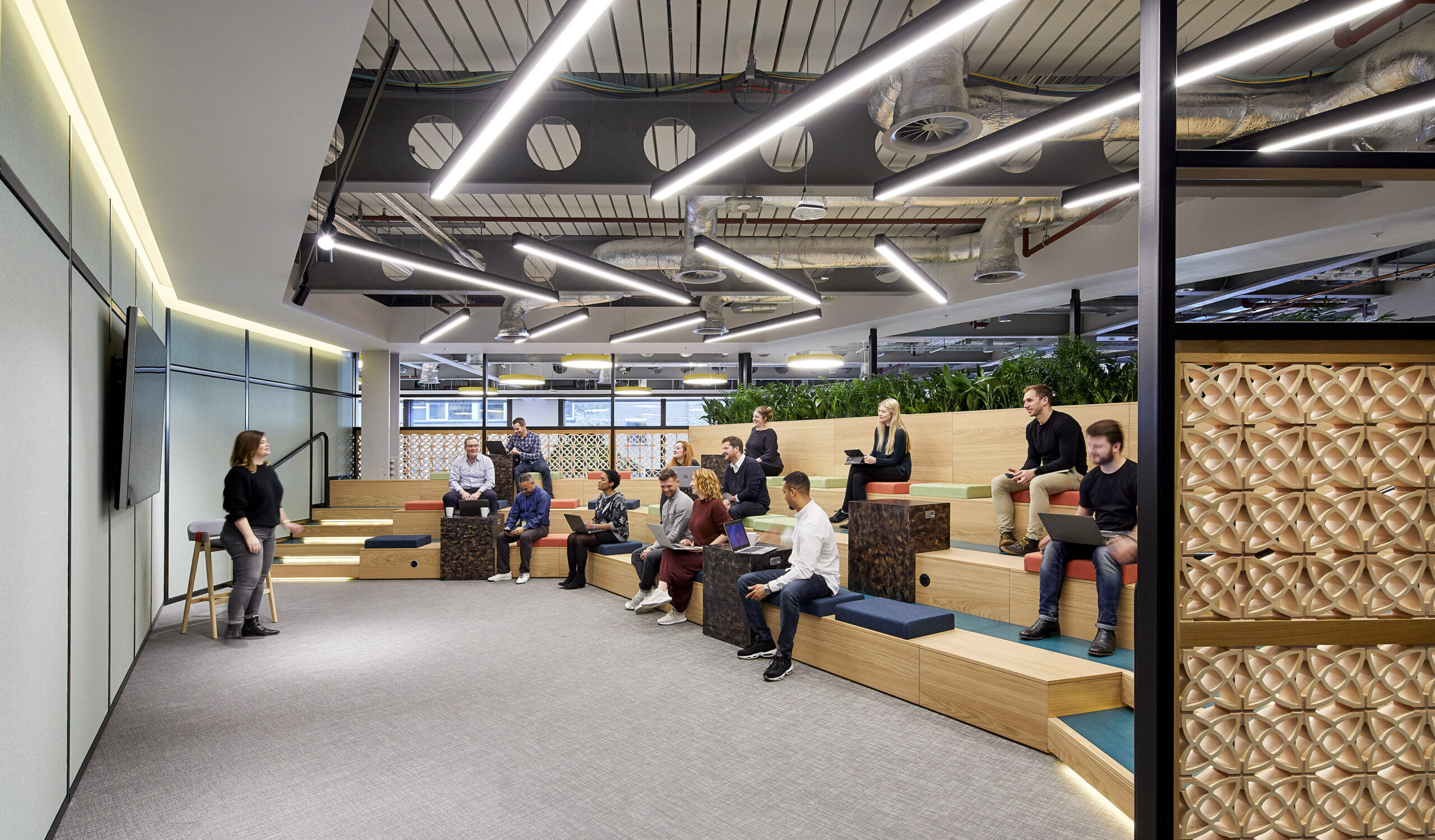 Modern collaborative workspace featuring tiered wooden seating designed for group discussions and presentations. Ambient lighting and vibrant green plants enhance the environment, while textured wall panels add visual interest. Flexible seating options promote comfort and creativity, fostering an innovative atmosphere for teamwork and interaction.