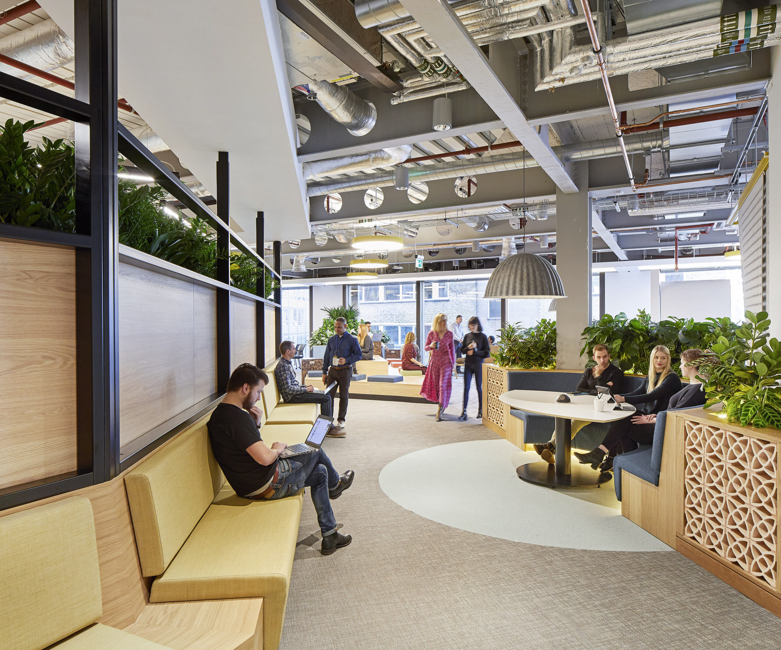 Brightly lit collaborative workspace featuring plush green seating and modern wooden accents. Lush indoor plants create a vibrant atmosphere, enhancing the biophilic design. The layout encourages interaction, with a communal table at the center, surrounded by comfortable seating areas, promoting teamwork and innovation within the office environment.