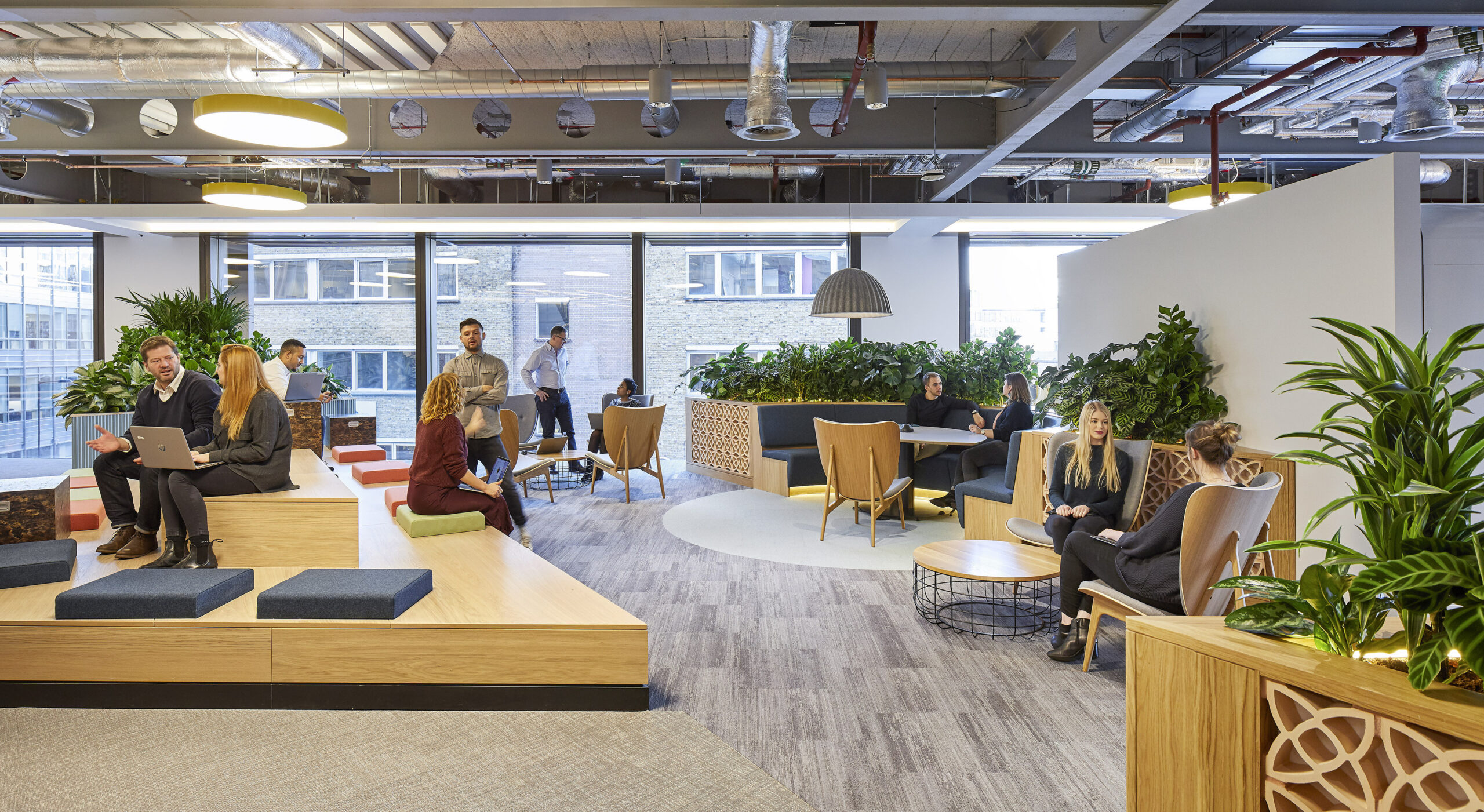 A modern office interior featuring a variety of seating areas, including plush chairs and tiered wooden platforms. Abundant greenery enhances the space, while large windows provide natural light. Flexible workstations accommodate collaboration, promoting a vibrant and innovative atmosphere conducive to creativity and productivity.