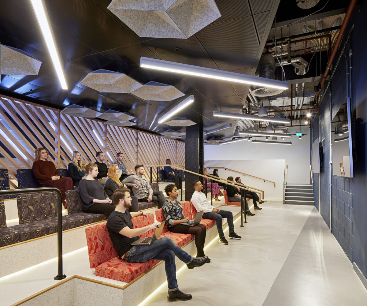 A modern auditorium featuring tiered seating with vibrant, patterned cushions. Attendees are engaged, facing a large screen. The ceiling is adorned with hexagonal acoustic panels and sleek linear lighting, creating a dynamic yet inviting atmosphere. Wood paneling along the walls enhances the contemporary aesthetic.