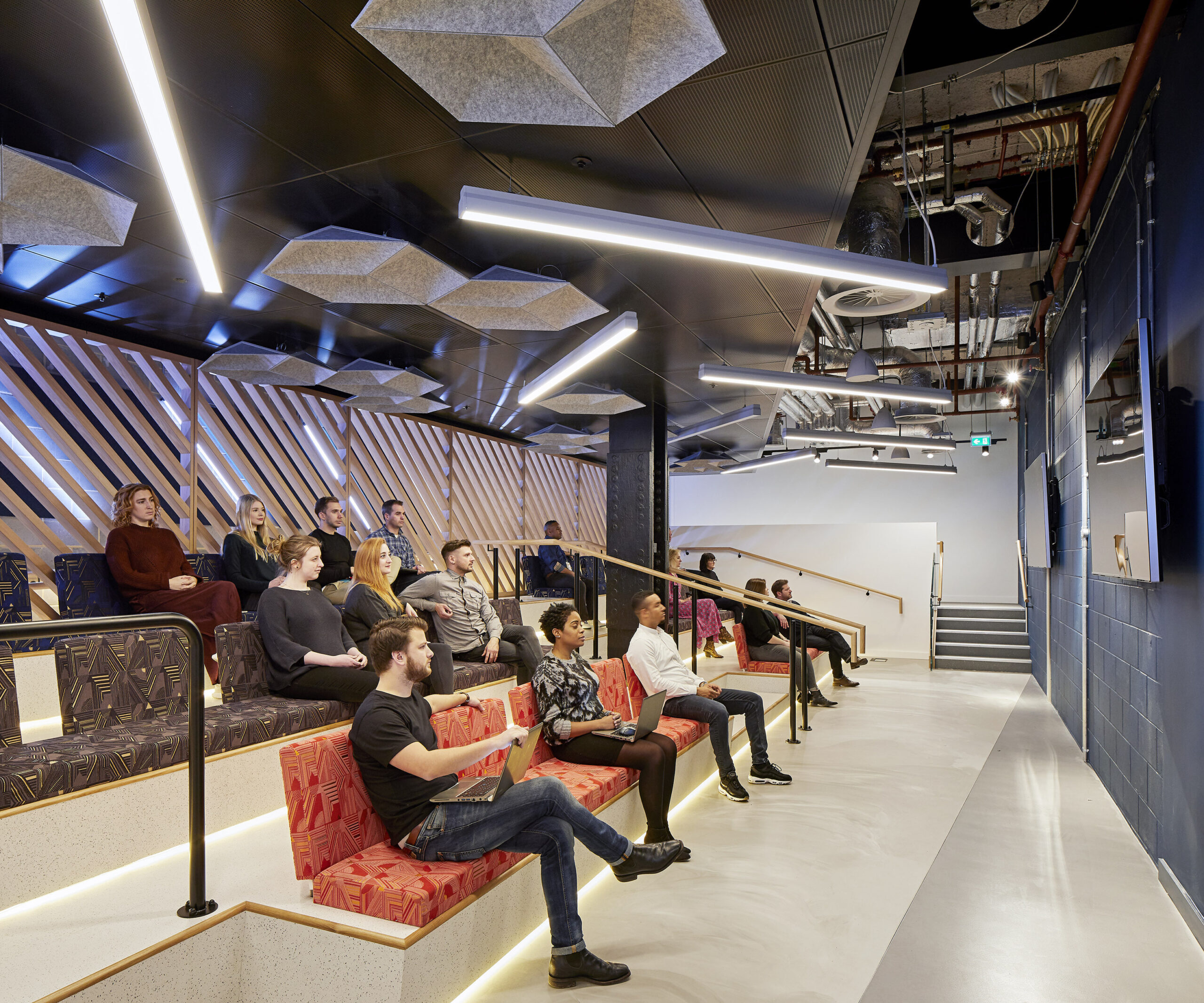A modern auditorium featuring tiered seating with vibrant, patterned cushions. Attendees are engaged, facing a large screen. The ceiling is adorned with hexagonal acoustic panels and sleek linear lighting, creating a dynamic yet inviting atmosphere. Wood paneling along the walls enhances the contemporary aesthetic.