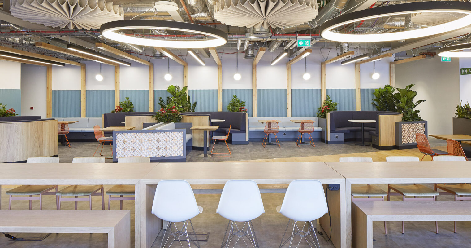 Bright and airy coworking space featuring large circular light fixtures and soft acoustic panels on the ceiling. The space utilizes a mix of wood and modern furniture, complemented by greenery. Flexible seating arrangements promote collaboration, while muted colors create a calm, inviting atmosphere for productivity.