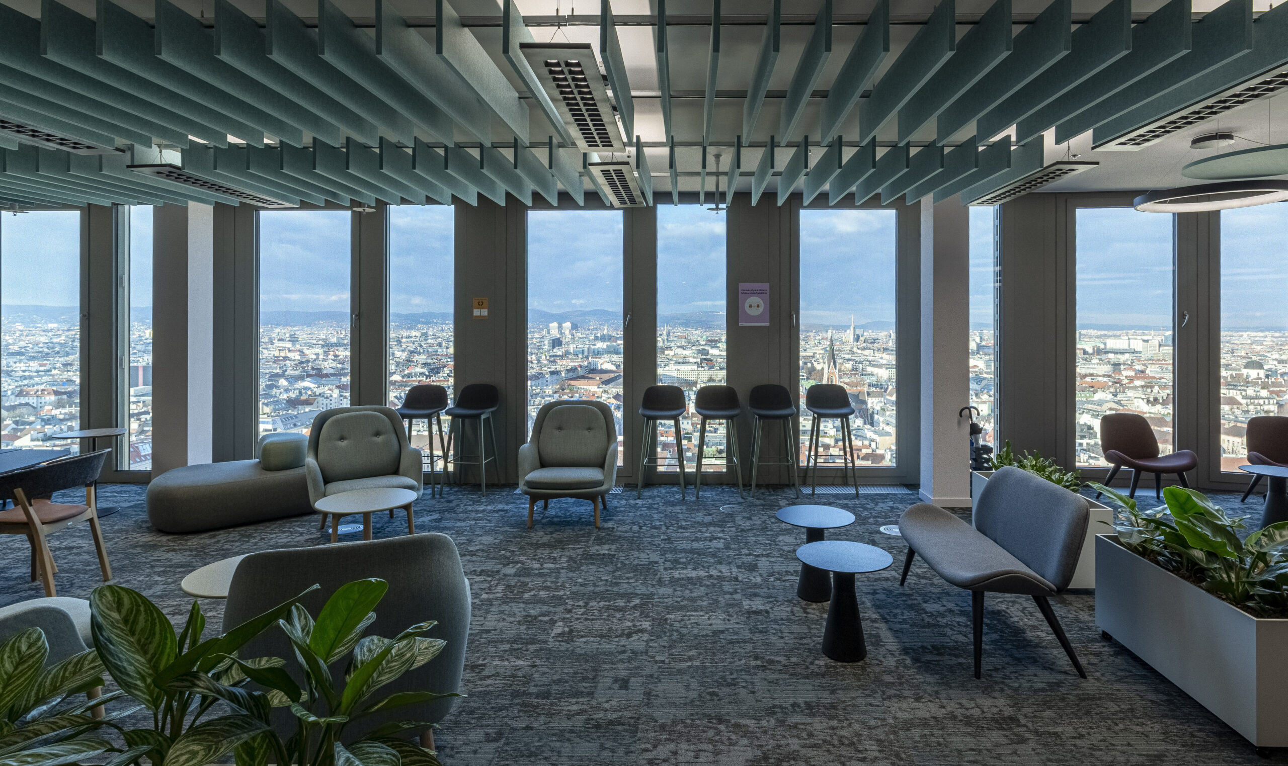 A spacious modern office lounge featuring panoramic city views through large windows. Stylish seating options, including upholstered chairs and sleek sofas, surround minimalist coffee tables. Acoustic paneling along the ceiling enhances sound quality, while strategically placed greenery adds a refreshing touch to the contemporary space.