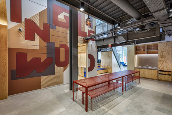A modern interior featuring an open layout with a bold mural in warm tones, incorporating geometric patterns. The space includes a sleek red metal table and minimalist wooden cabinetry, emphasizing a blend of industrial and contemporary design elements. Ample natural light highlights the innovative use of materials.