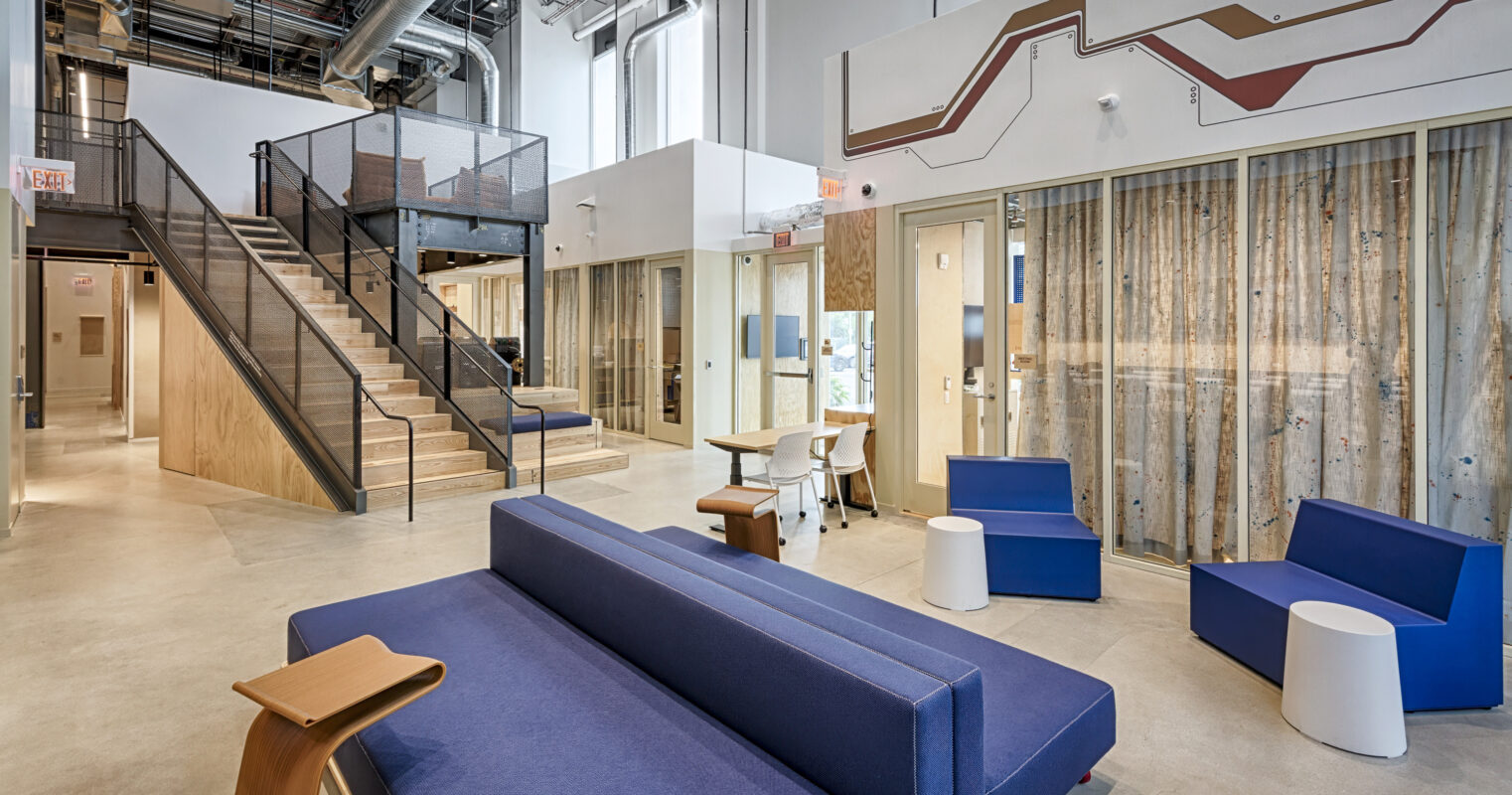 A spacious modern interior features a mix of wooden and industrial elements. Light floods the area through large windows, highlighting the combination of blue seating and sleek, minimalist furniture. An open staircase leads to upper levels, while textured wooden partitions offer privacy without compromising the airy ambiance.