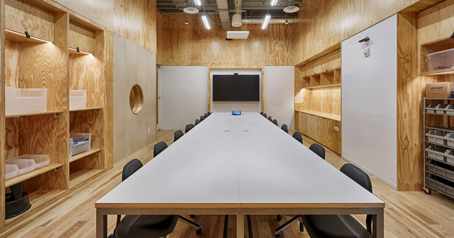 A spacious conference room features a long, minimalistic table surrounded by black chairs. The warm wood paneling creates a welcoming atmosphere, complemented by strategically placed shelves for storage. A large screen at the front and circular cutouts in the walls enhance both functionality and aesthetic appeal.