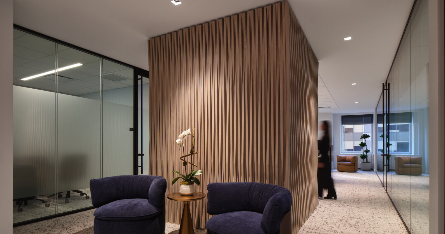 Luxurious office corridor showcasing a striking wood-paneled feature wall with vertical ridges, creating texture and warmth. Plush navy armchairs are paired with a sleek round coffee table, enhancing the inviting atmosphere. Glass partitions on either side allow light to flow, promoting an open yet professional environment.