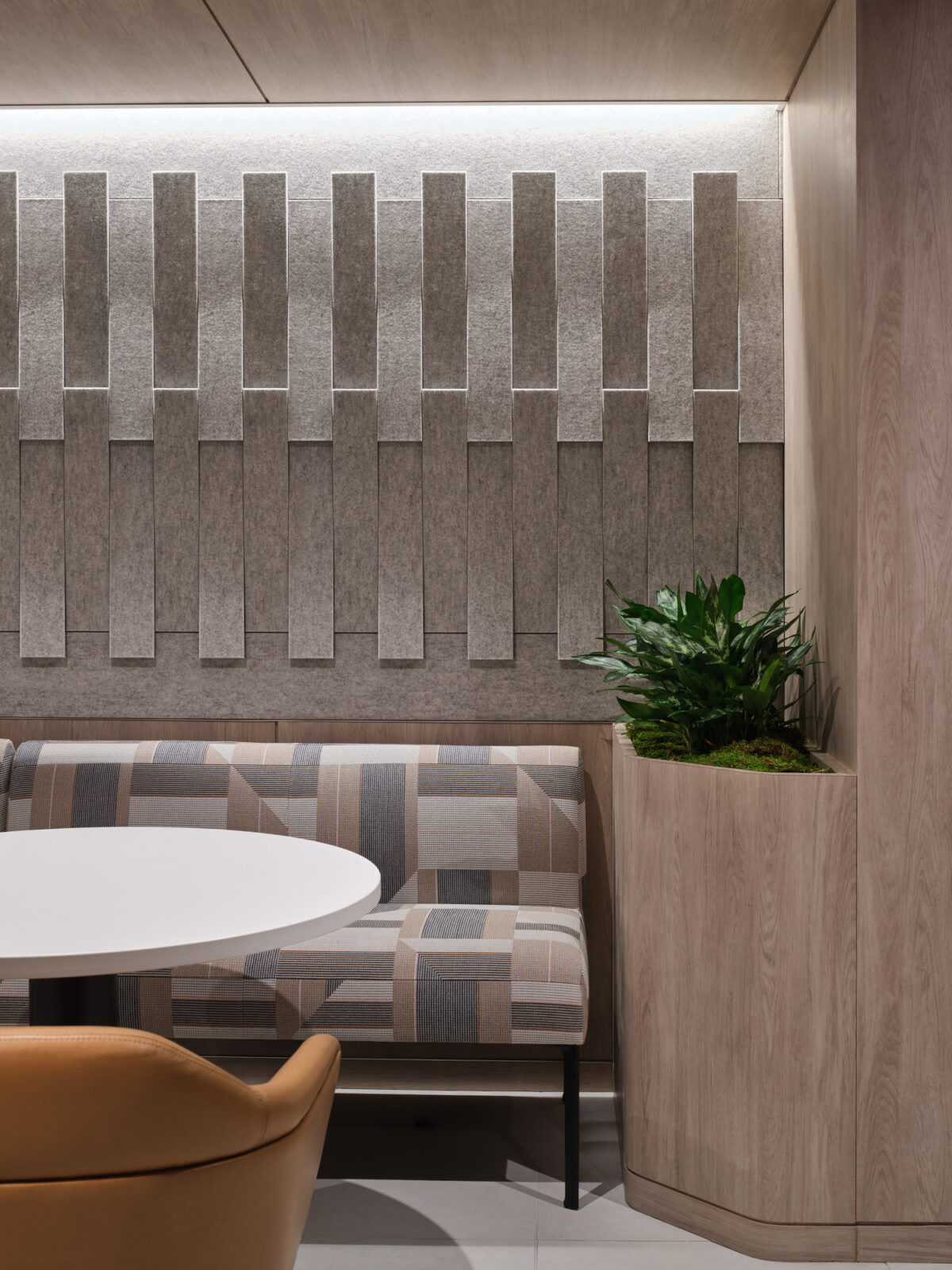 A modern seating area featuring a textured gray wall with vertical acoustic panels. A round white table is complemented by a cozy, patterned bench, while a sleek wooden planter housing lush greenery adds a touch of nature, creating a tranquil and inviting atmosphere perfect for collaborative spaces.