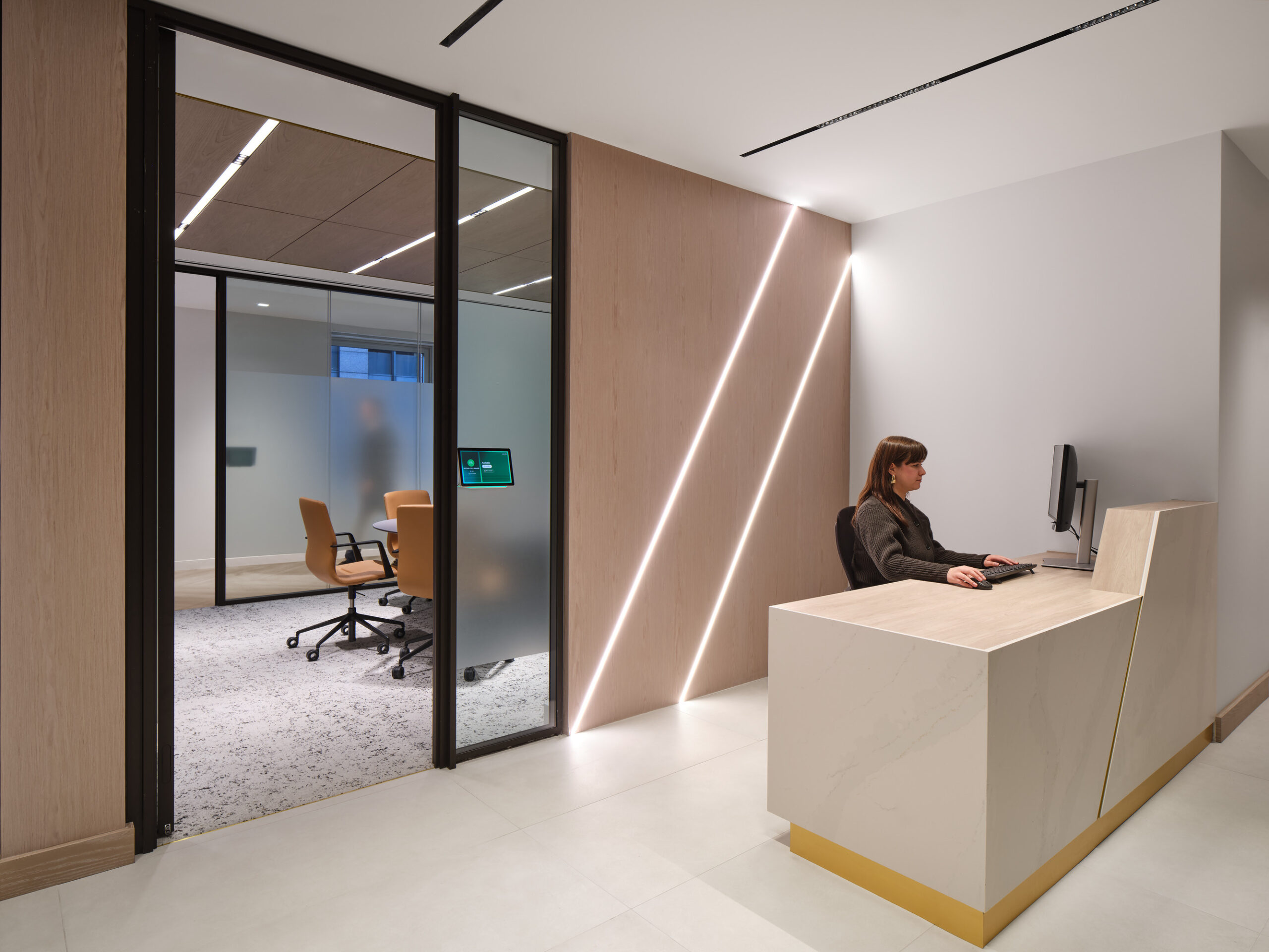 A modern office reception area features a sleek, minimalist wooden reception desk with a polished stone surface. Behind the desk, a professional woman is engaged with her computer. Glass sliding doors lead to a conference room with contemporary seating and a light color palette, illuminated by subtle linear lighting.