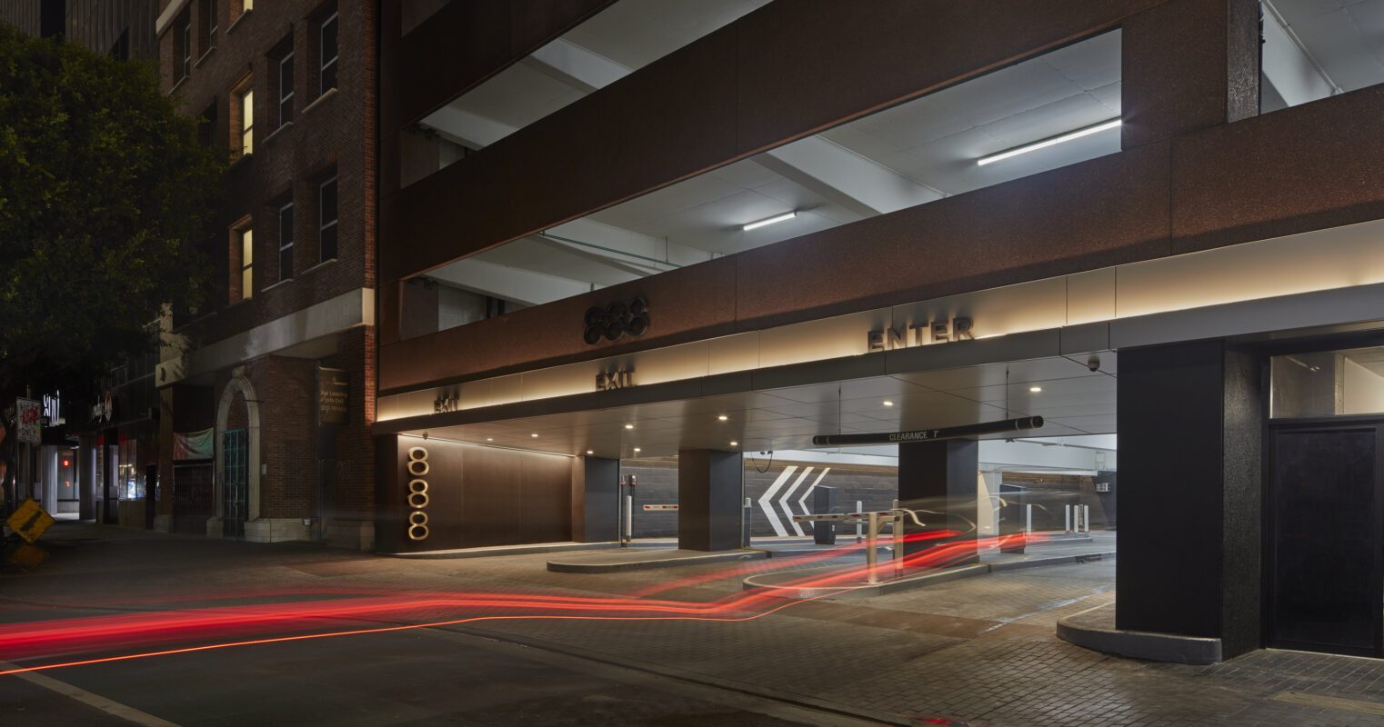 The entrance to a modern parking garage features sleek, dark facade materials contrasted by illuminated signage. Architectural lines guide vehicles with bold arrows, while subtle ambient lighting enhances safety and aesthetic appeal. Nearby brick buildings create a harmonious blend between contemporary design and historical architecture.