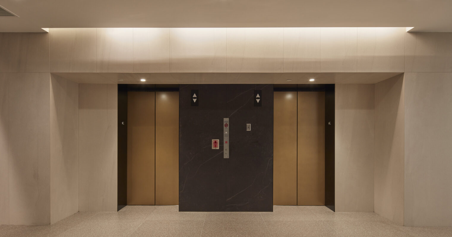 The elevator area features a minimalist design with a sleek marble accent wall in dark tones, complemented by brass elevator doors. Soft, recessed lighting enhances the contemporary aesthetic, while the polished concrete flooring introduces texture and sophistication, creating an inviting yet modern entrance to the building.
