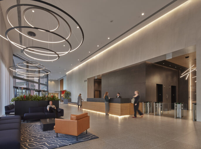 Modern lobby featuring sleek, minimalist design with high ceilings and natural light. Suspended circular LED light fixtures create a focal point above the reception area. A large, textured wall complements the contemporary furnishings, including plush sofas and a distinctive orange chair. Lush greenery adds warmth to the space.