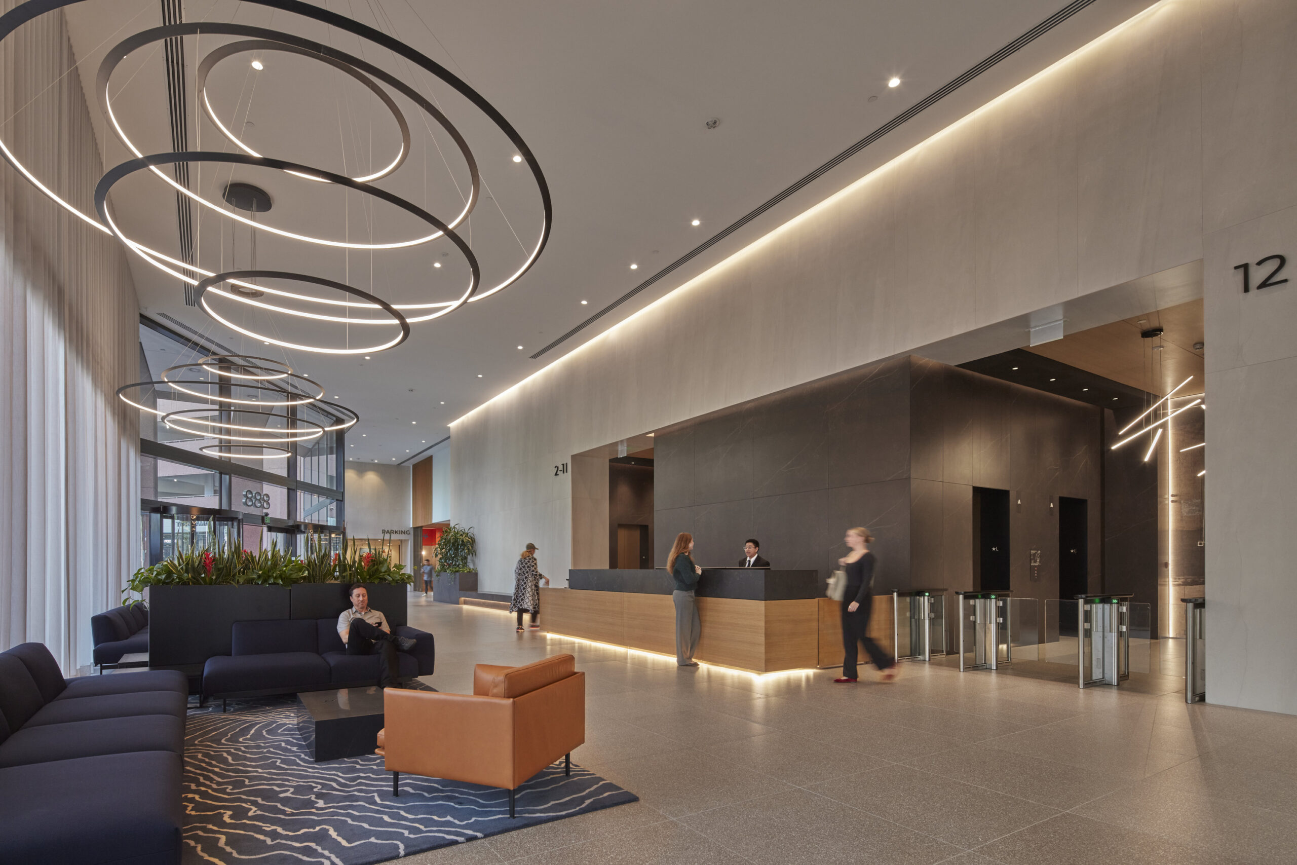 Modern lobby featuring sleek, minimalist design with high ceilings and natural light. Suspended circular LED light fixtures create a focal point above the reception area. A large, textured wall complements the contemporary furnishings, including plush sofas and a distinctive orange chair. Lush greenery adds warmth to the space.
