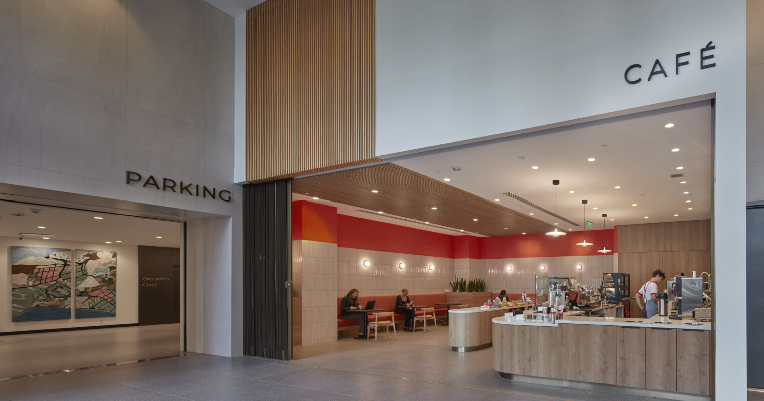 Bright and welcoming lobby featuring a café and parking entrance. The space is defined by smooth stone flooring, wooden accent walls, and contemporary artwork. The café area showcases a sleek counter with bar stools, inviting seating, and vibrant red accents, creating a warm and friendly atmosphere.