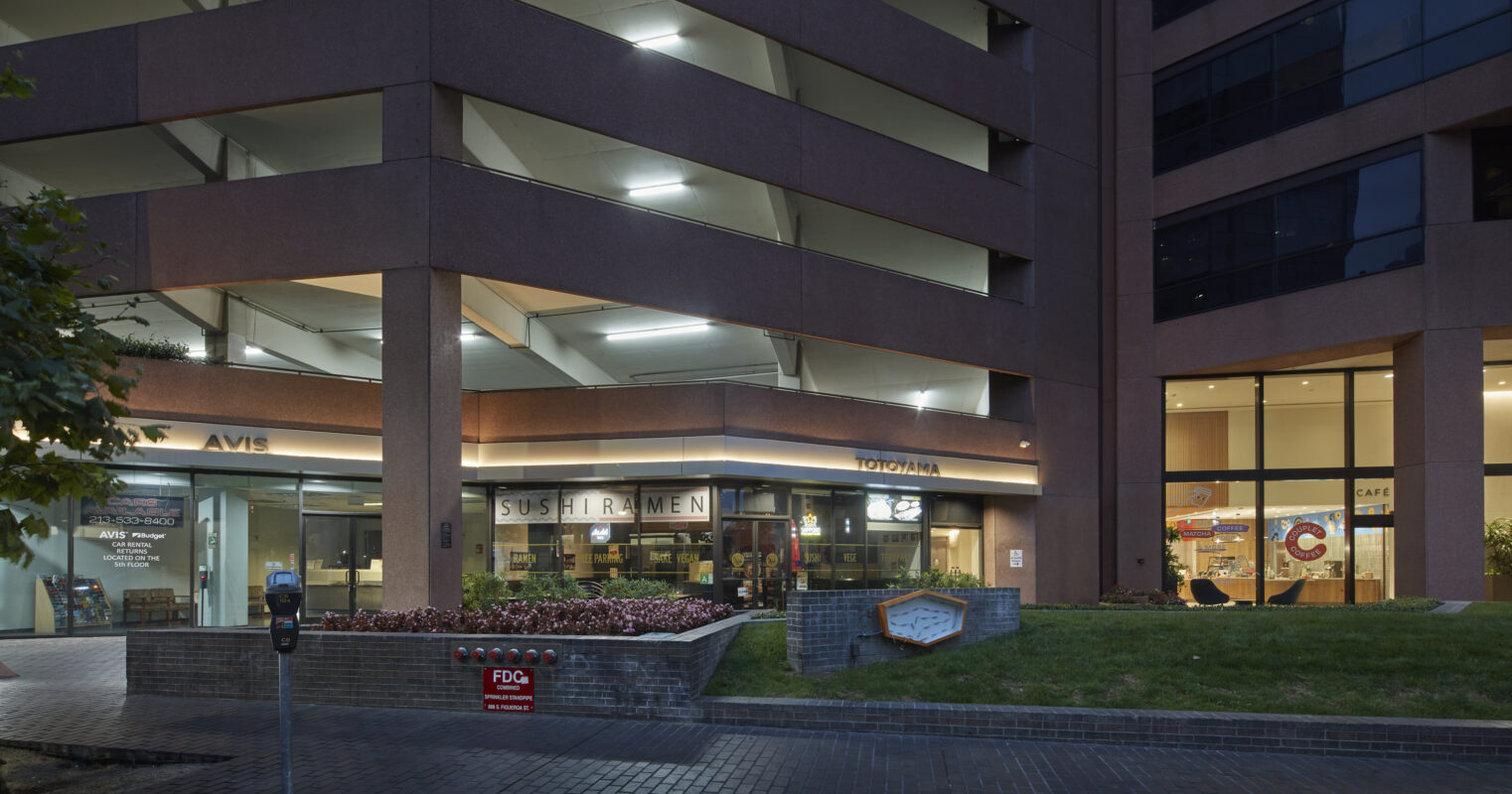 The entrance of a modern mixed-use building features a welcoming atrium with large glass windows showcasing vibrant restaurants and cafés. Lush landscaping with colorful flower beds and pathway lighting enhances the outdoor space, creating an inviting atmosphere for both pedestrians and patrons. Compact parking meters are strategically placed for convenience.